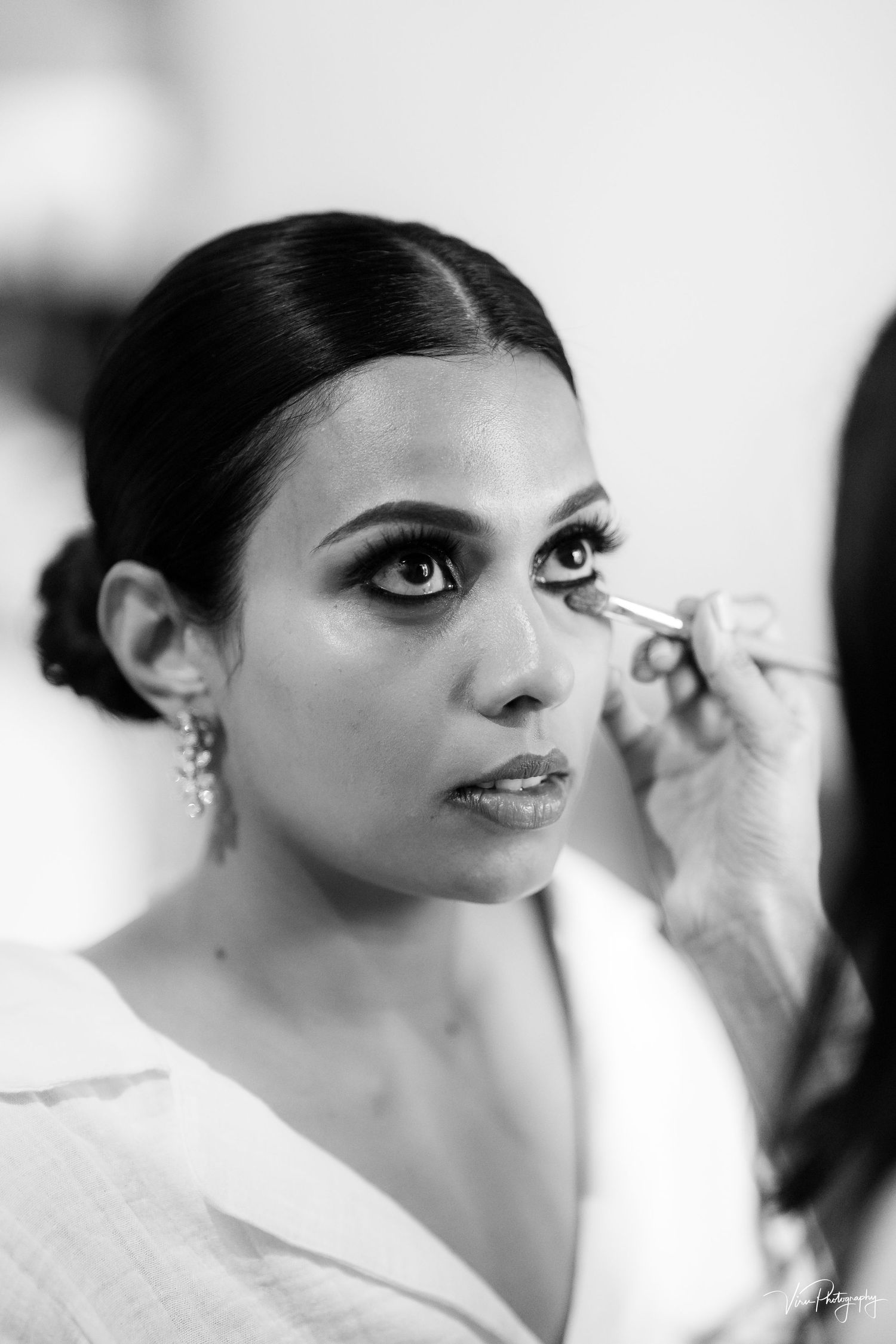 Black and white portrait shows preparations with makeup application, focusing on glamorous eye details and elegant earrings.