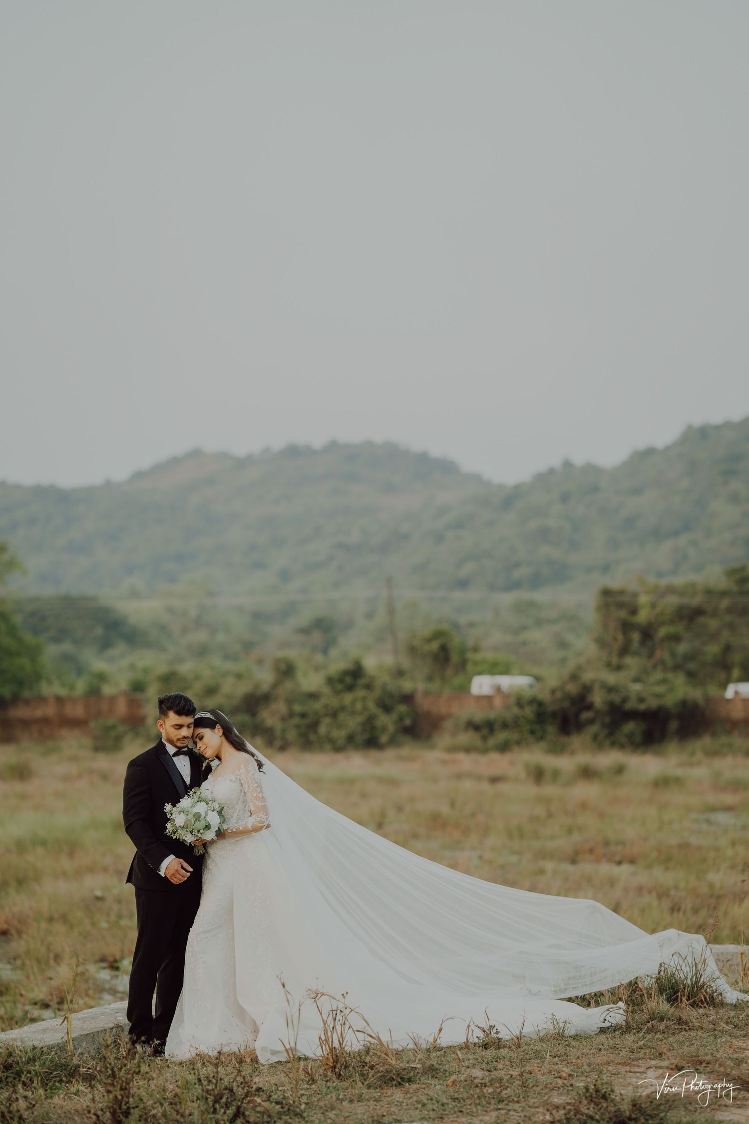 Couple stands in scenic mountainous field during romantic wedding photoshoot with flowing white dress and lush greenery.