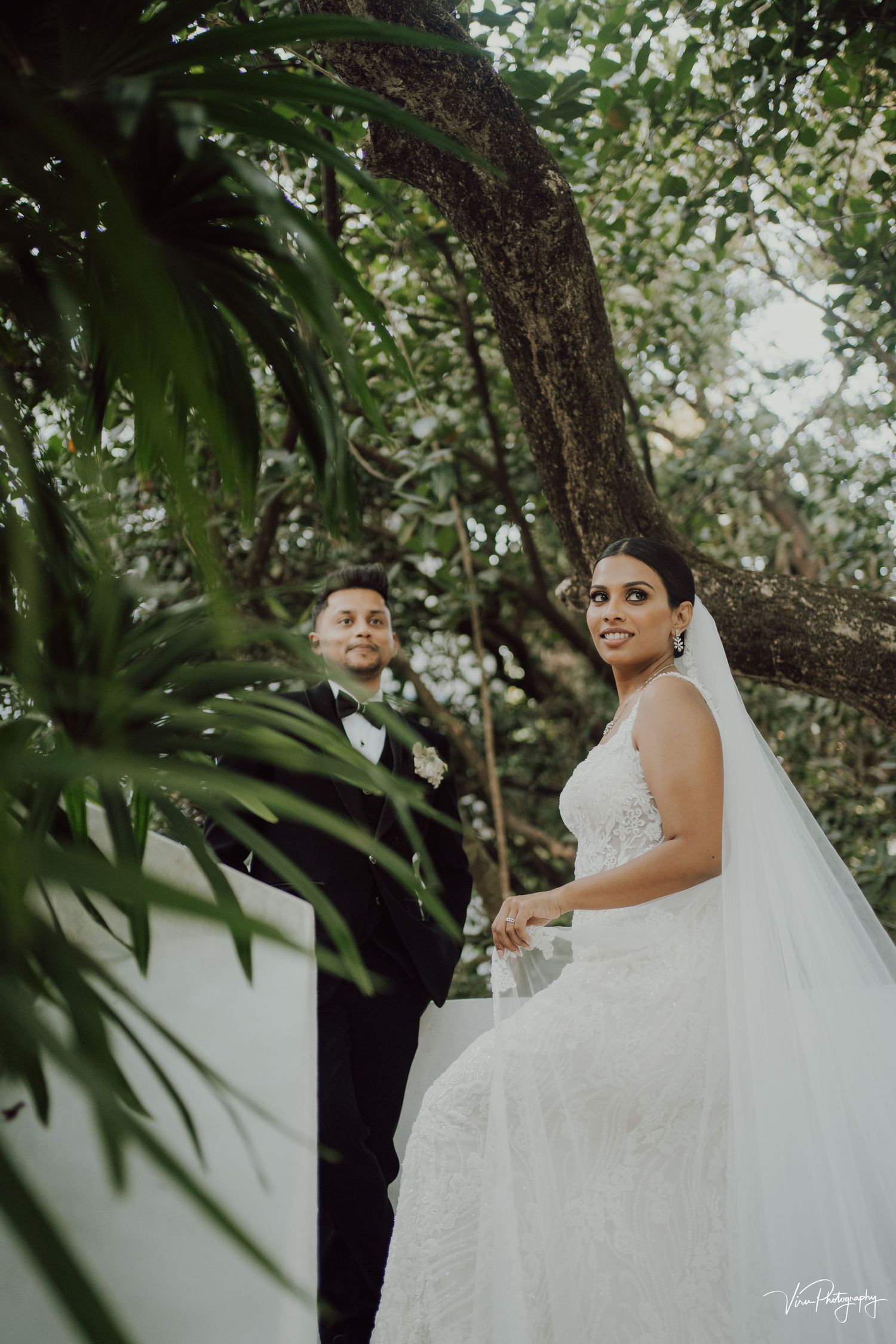 A couple shares a romantic moment among lush greenery and trees during an outdoor wedding ceremony.
