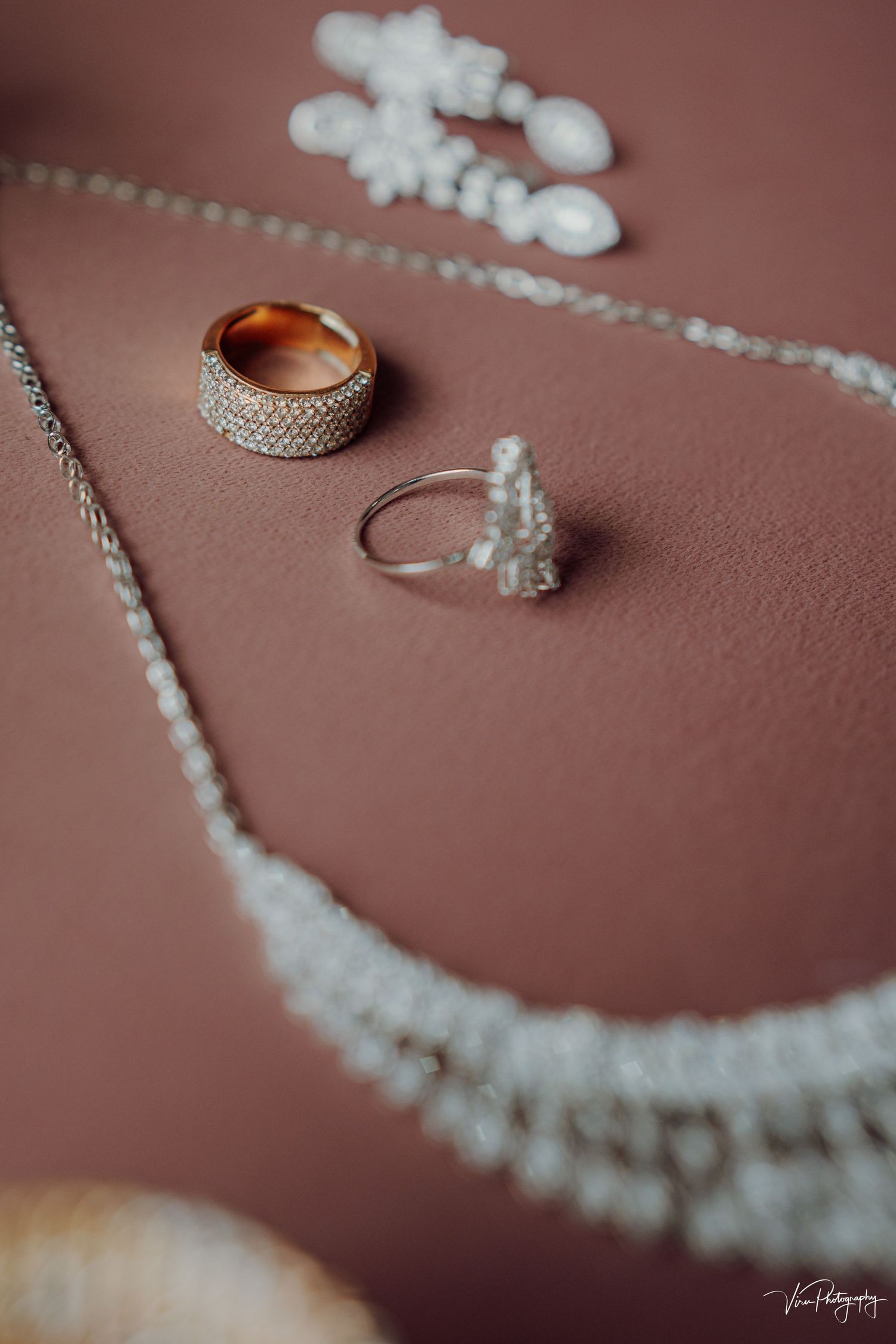 Wedding jewelry including a diamond encrusted band, delicate ring, and pearl necklace arranged on pink surface.