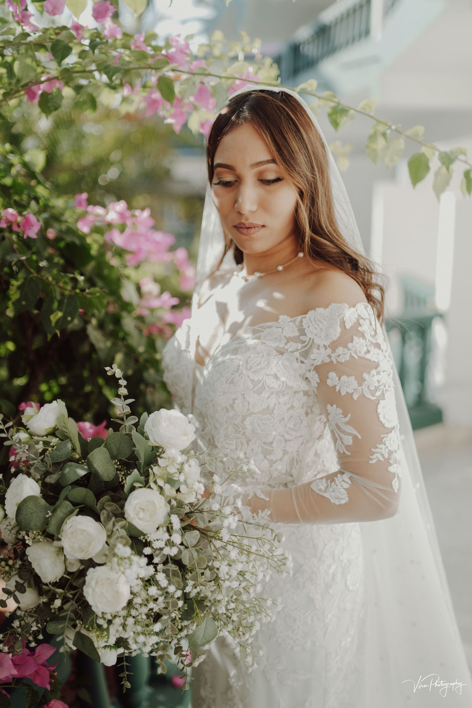 A bride in a delicate lace wedding dress with sheer sleeves holds an elegant white rose bouquet amid pink flowering vines.