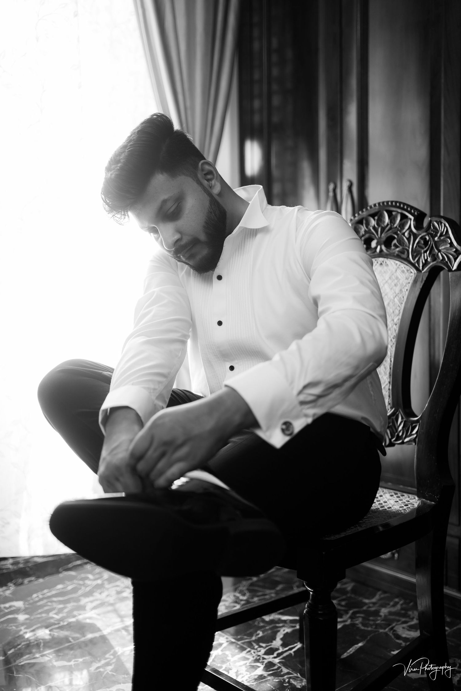 Black and white portrait of a person in a white dress shirt and dark pants sitting thoughtfully in an ornate chair.
