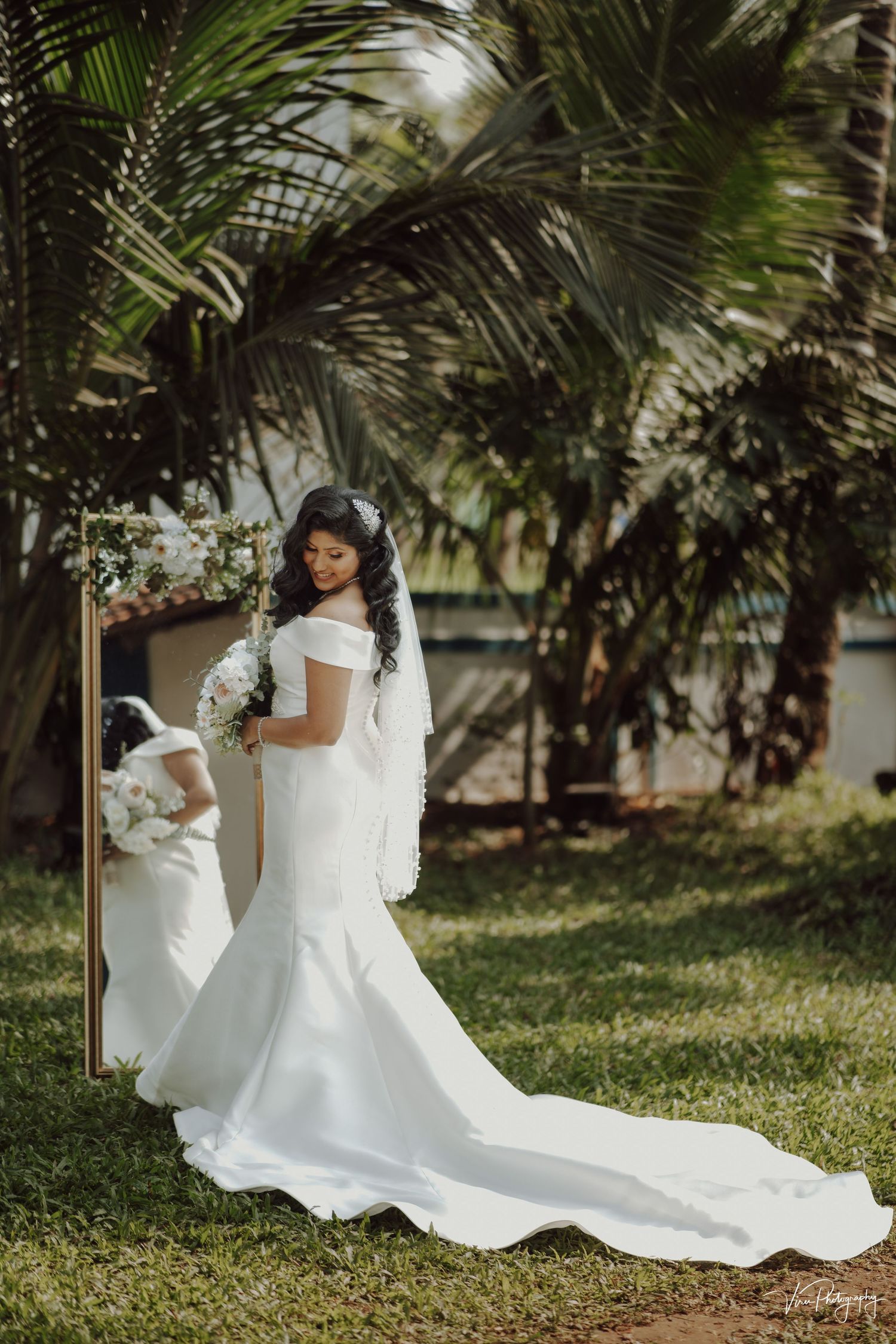 A bride in an elegant white mermaid wedding dress with a long train poses outdoors among tropical palm trees.