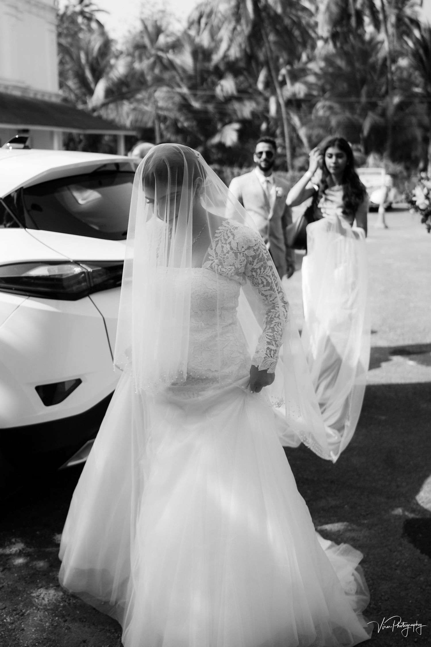 Black and white photo of a bridal party outside with palm trees, featuring elegant wedding dresses and a white car.