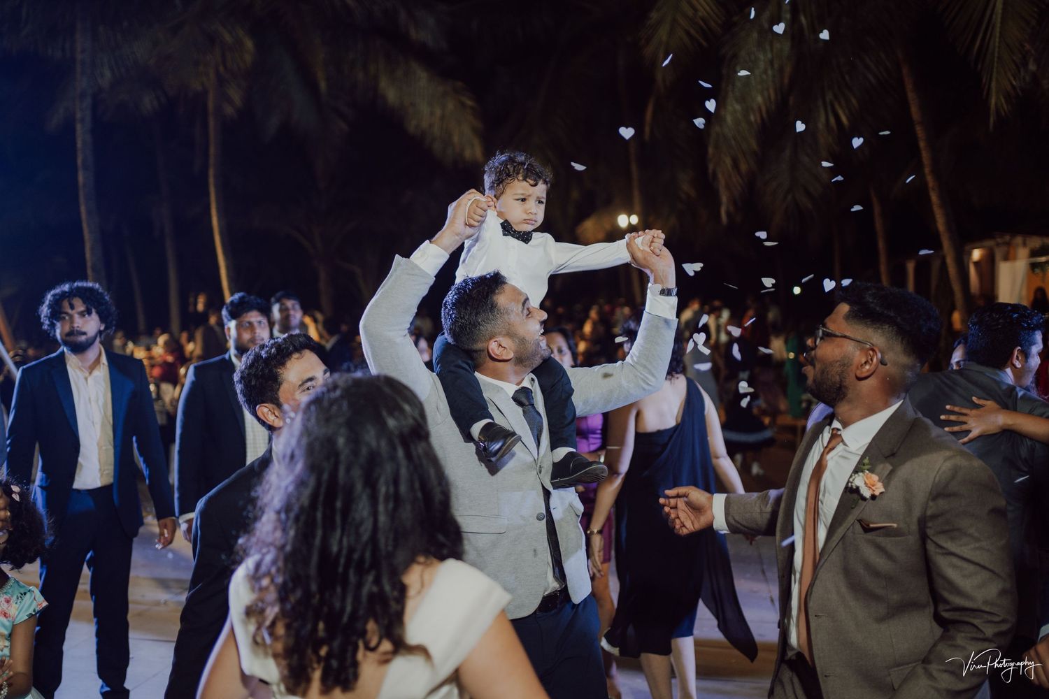 Joyful guests celebrate on a dance floor at an outdoor nighttime wedding reception surrounded by palm trees and twinkling lights.