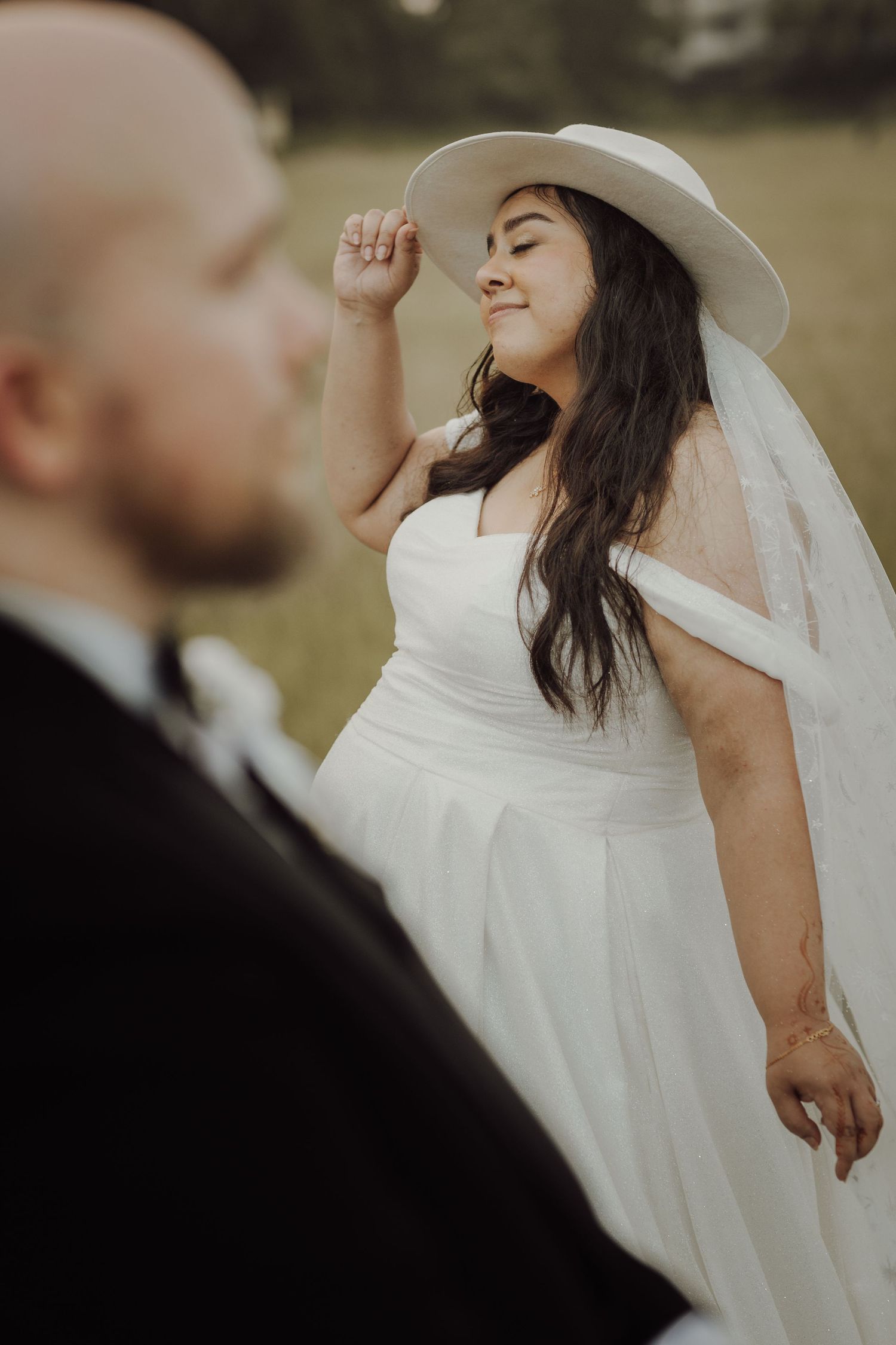 A bride in a flowing white dress and hat playfully tilts her head back during an outdoor wedding photoshoot.
