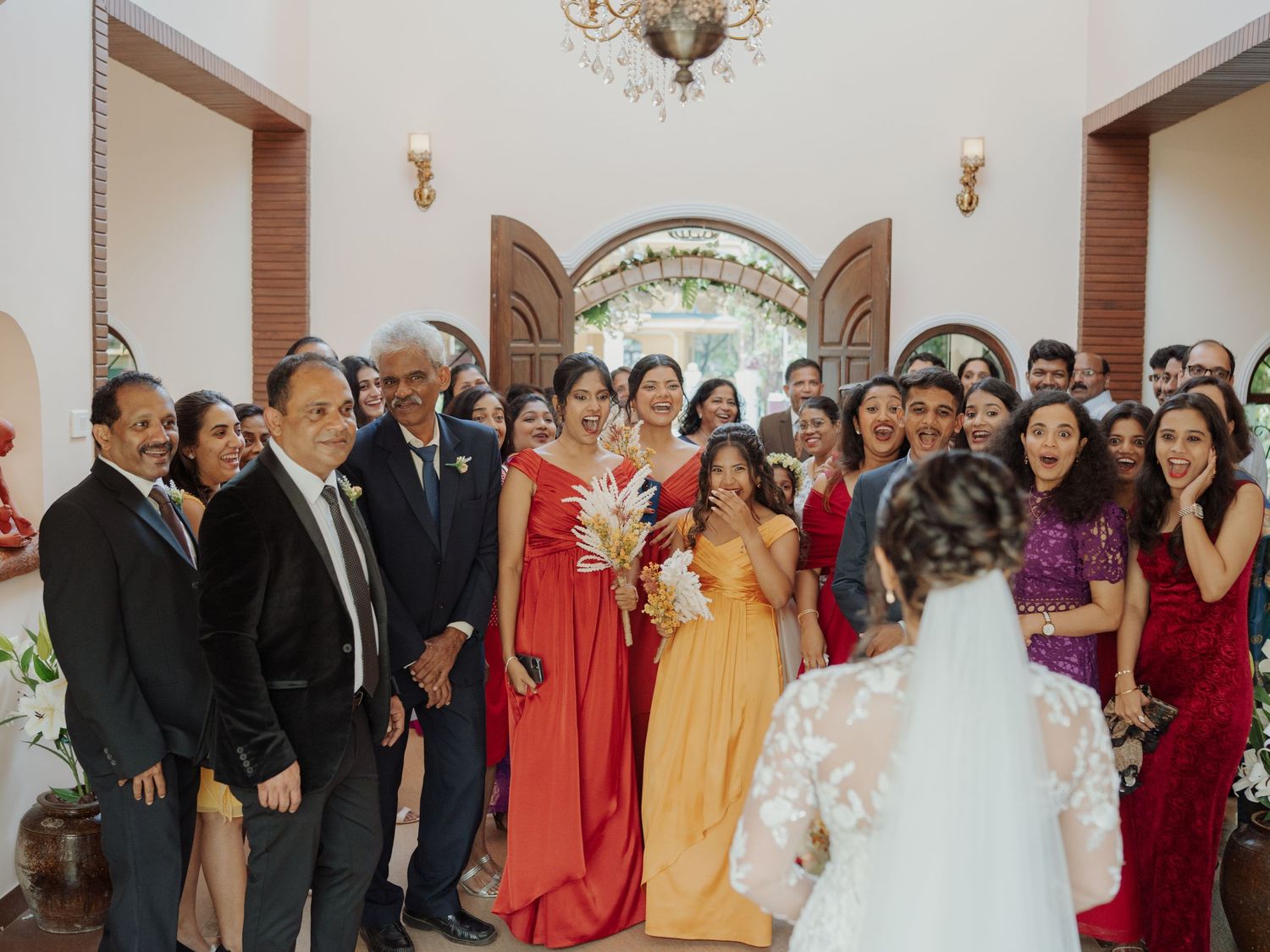 A bride in a white veil faces a smiling crowd of wedding guests in formal and red dresses inside a grand arched hall.