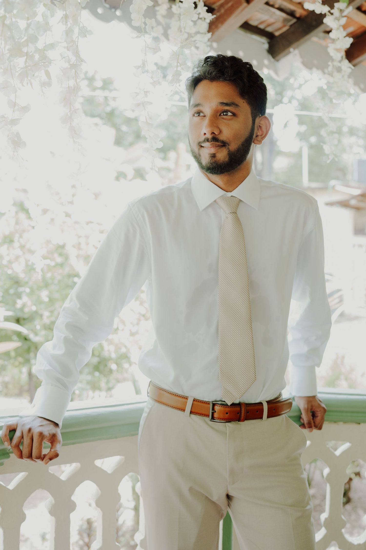A groom in a white shirt, cream tie and tan trousers leans on a balcony railing framed by greenery and white flowers.