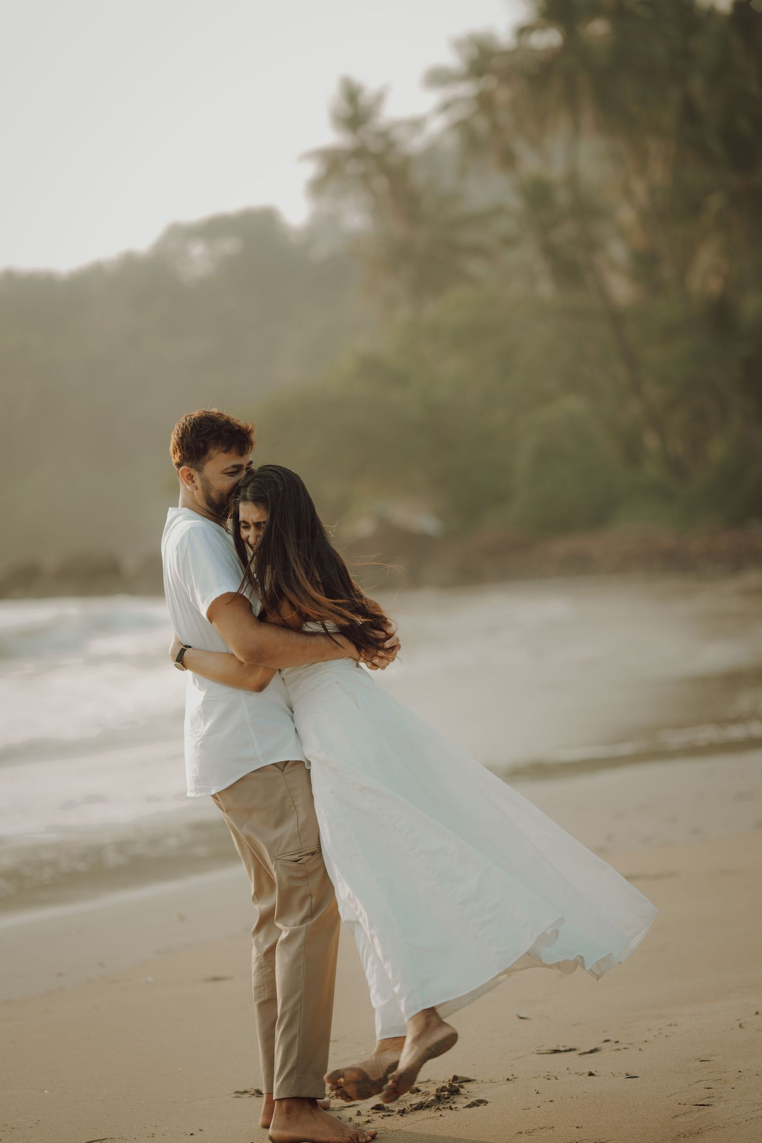 A couple embraces romantically on a misty tropical beach at sunset with palm trees in the background.