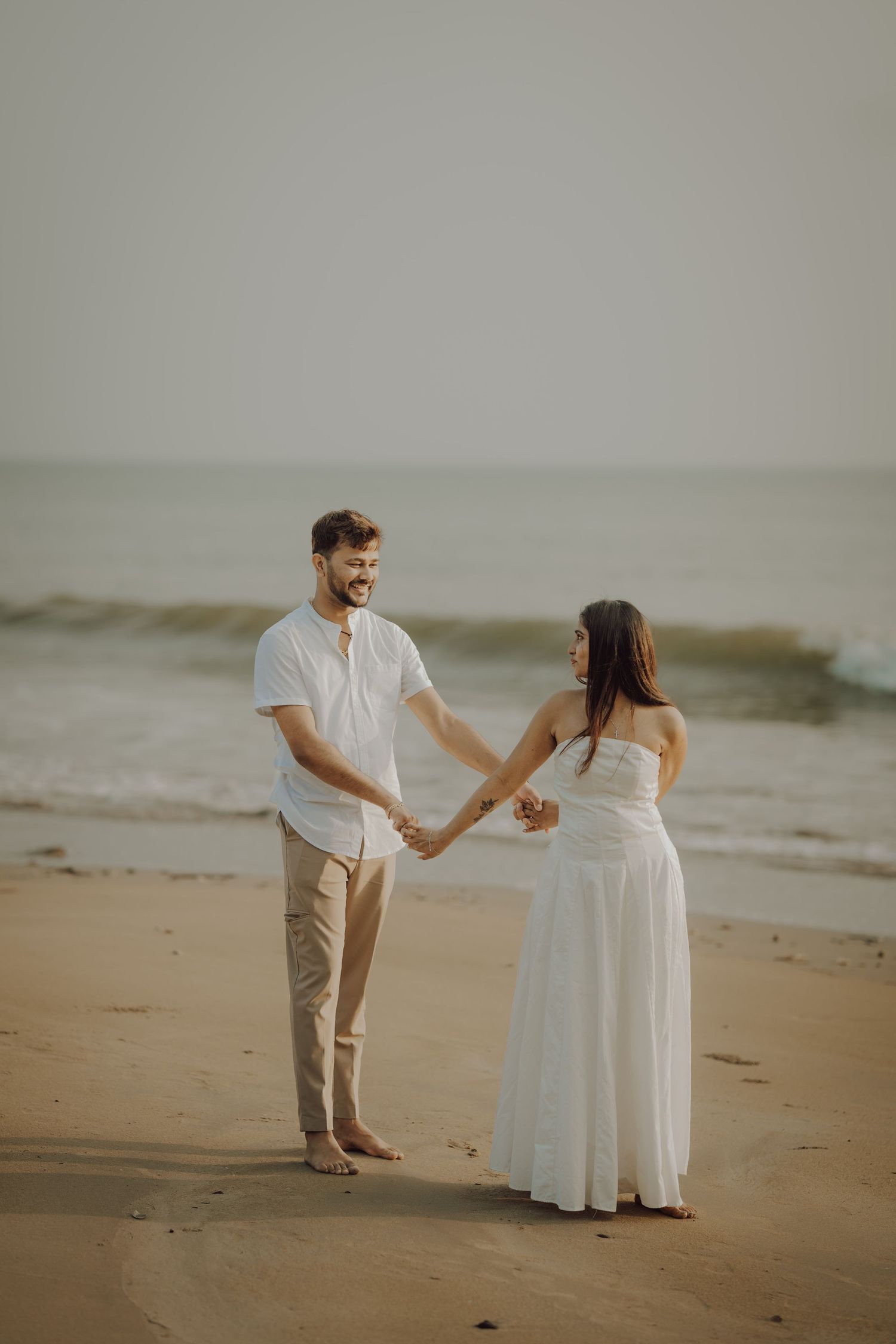 A couple in white and beige attire walks along a misty beach shore holding hands.
