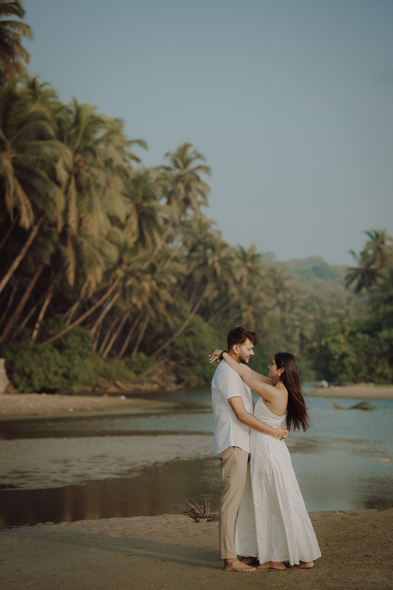 A couple in white outfits embrace romantically on a tropical beach with palm trees and mountains in the background.