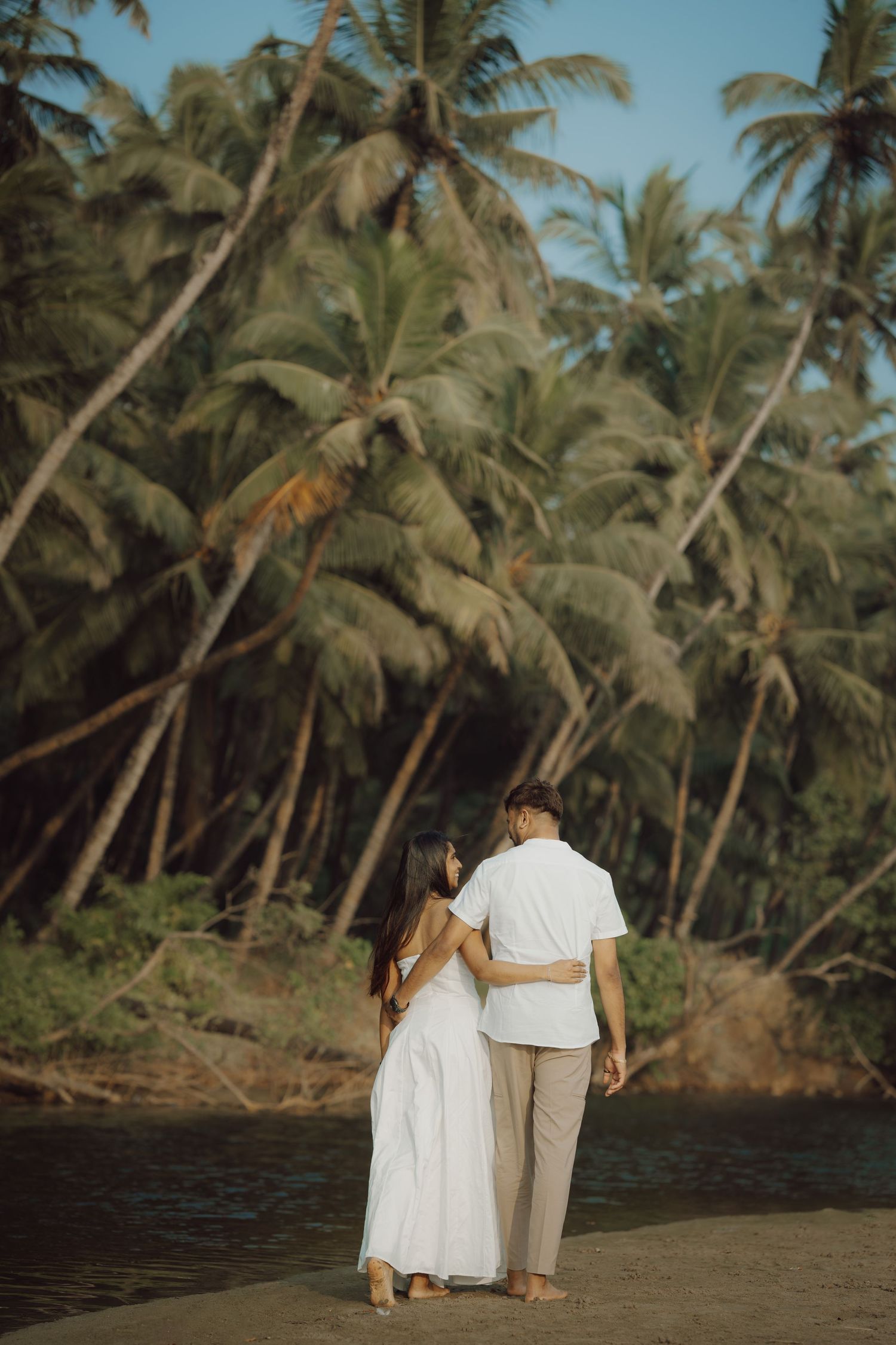 A couple walks together on a tropical beach surrounded by palm trees at sunset.