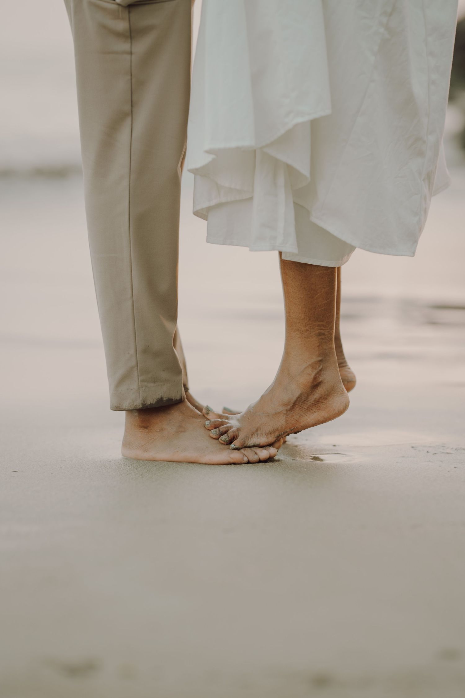 Close-up of bare feet on sand with one person wearing khaki pants and another in a flowing white dress.