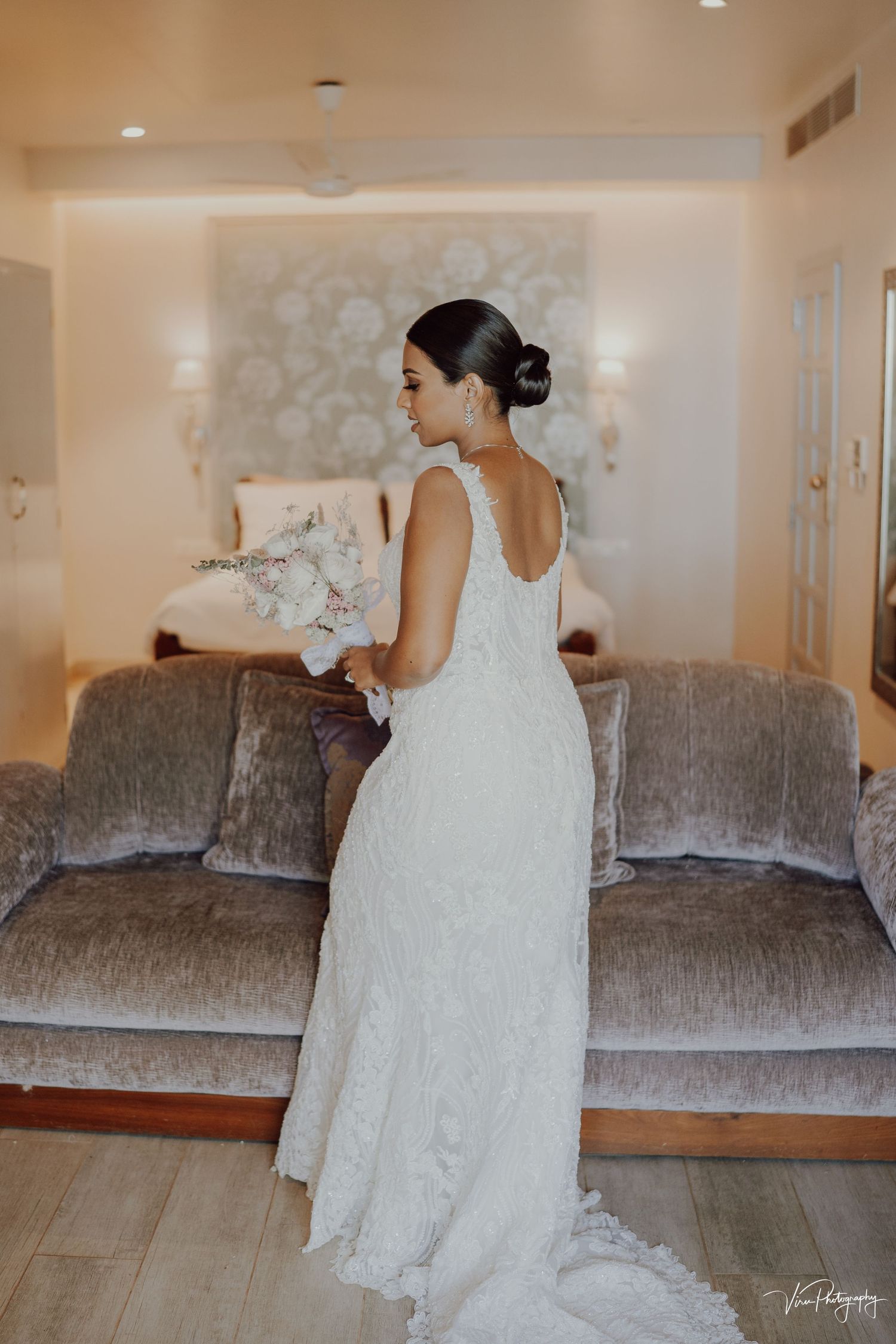 A bride in a white lace gown with an elegant updo hairstyle stands beside a gray velvet sofa on her wedding day.