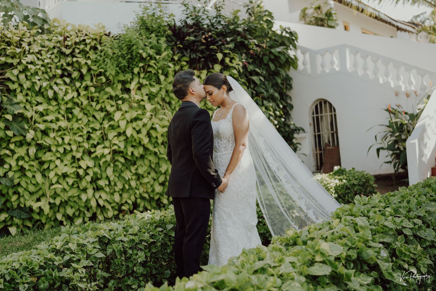 A newlywed couple embraces in a garden courtyard against a white wall and lush green foliage.
