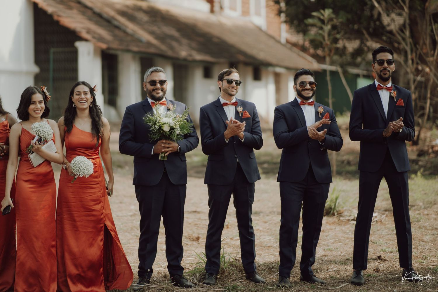 Wedding party in navy suits and red dresses pose together holding bouquets outside a rustic building.