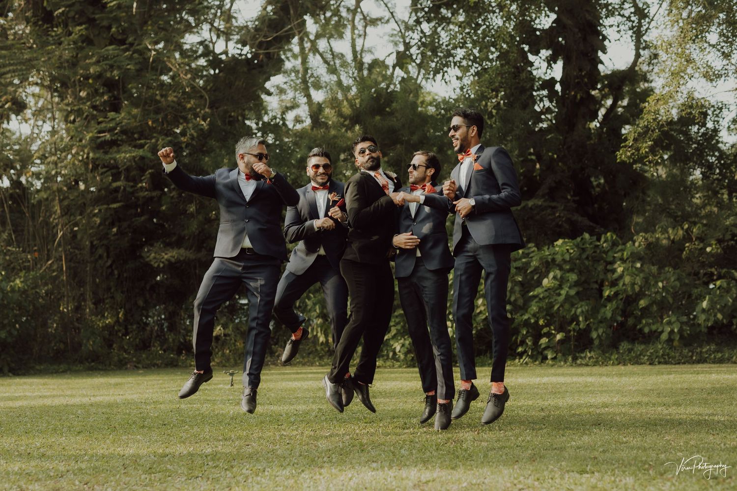 Joyful wedding party jumps together in unison on a green lawn surrounded by trees during an outdoor celebration.