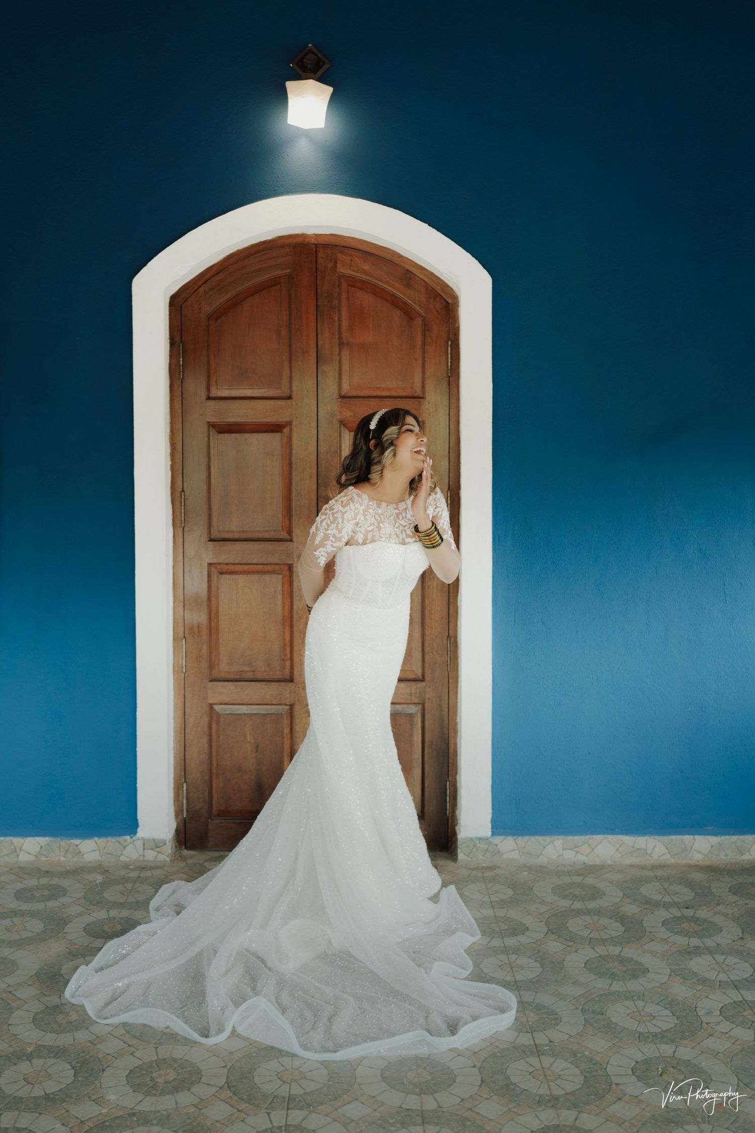 A bride in a flowing mermaid-style white wedding gown poses elegantly in front of a wooden arched doorway against a blue wall.