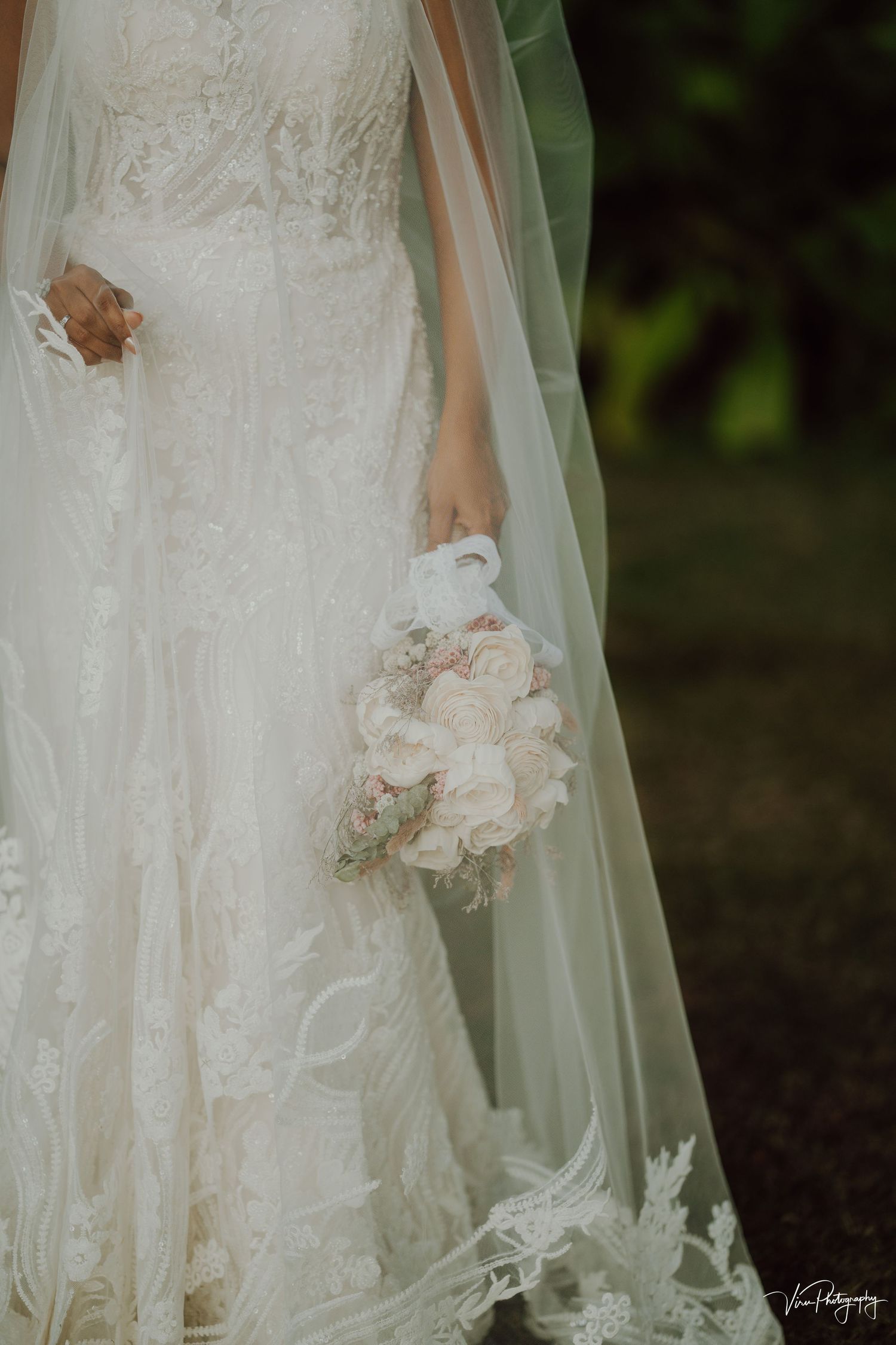 Close-up detail of a white lace wedding dress with soft floral bouquet against a dark background.