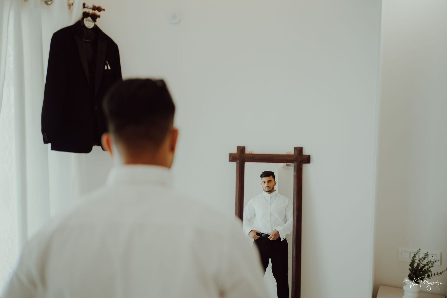 Person looks in mirror while getting ready with dark blazer hanging in minimalist white room setting.