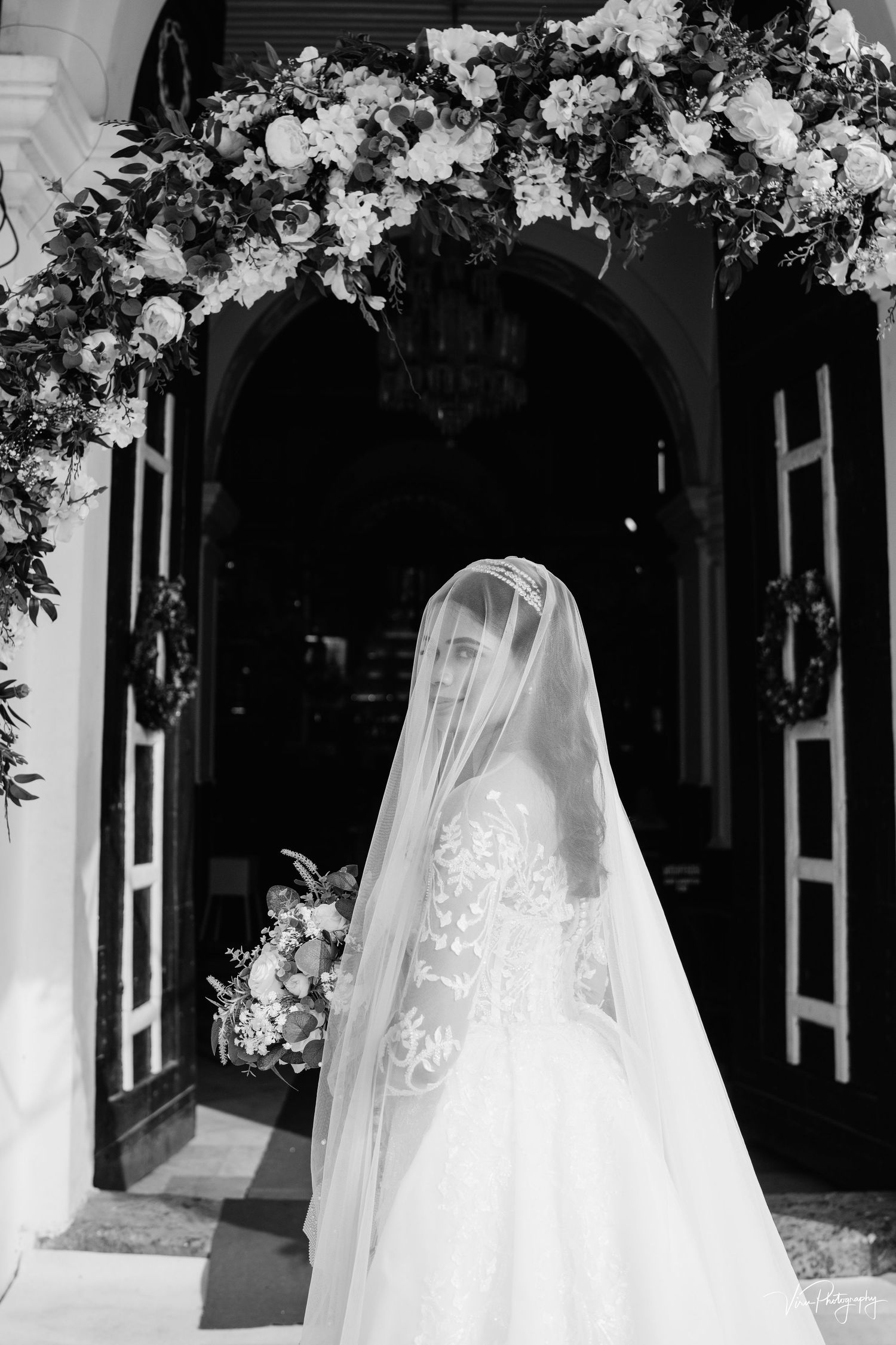 A bride in a white wedding gown and long veil stands in a church doorway adorned with white floral arrangements.