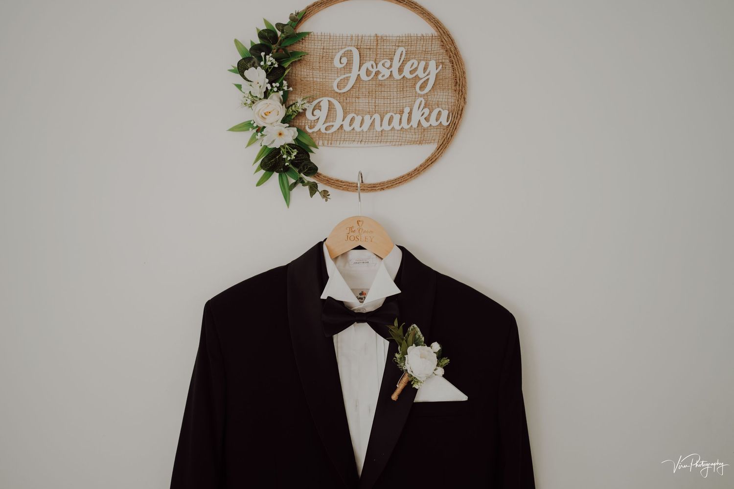 A dark wedding suit with bowtie hangs beneath a wooden circular sign decorated with greenery on a light gray wall.
