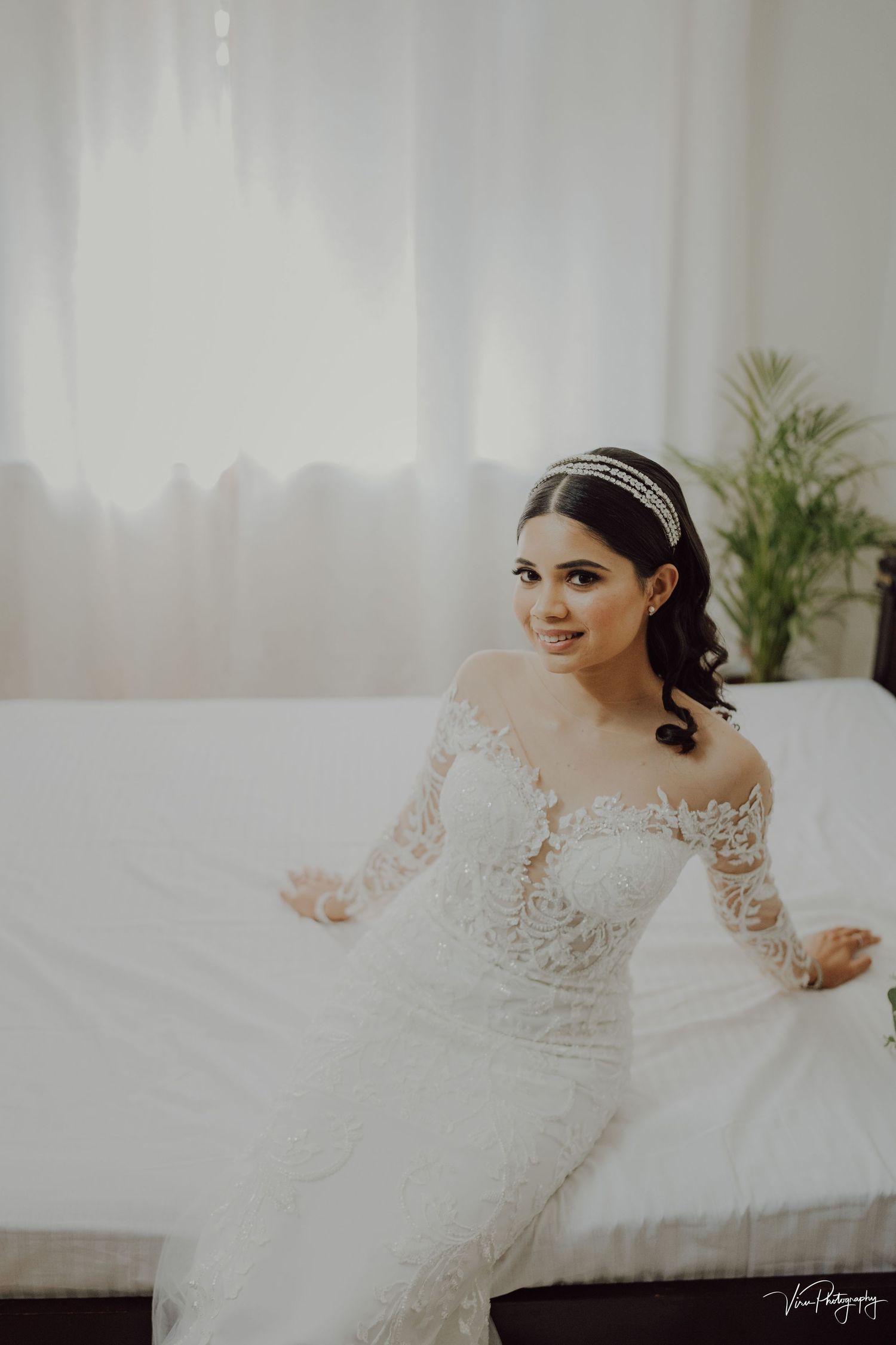 Bride in long-sleeve lace wedding dress and crystal headband poses gracefully in bright indoor setting.