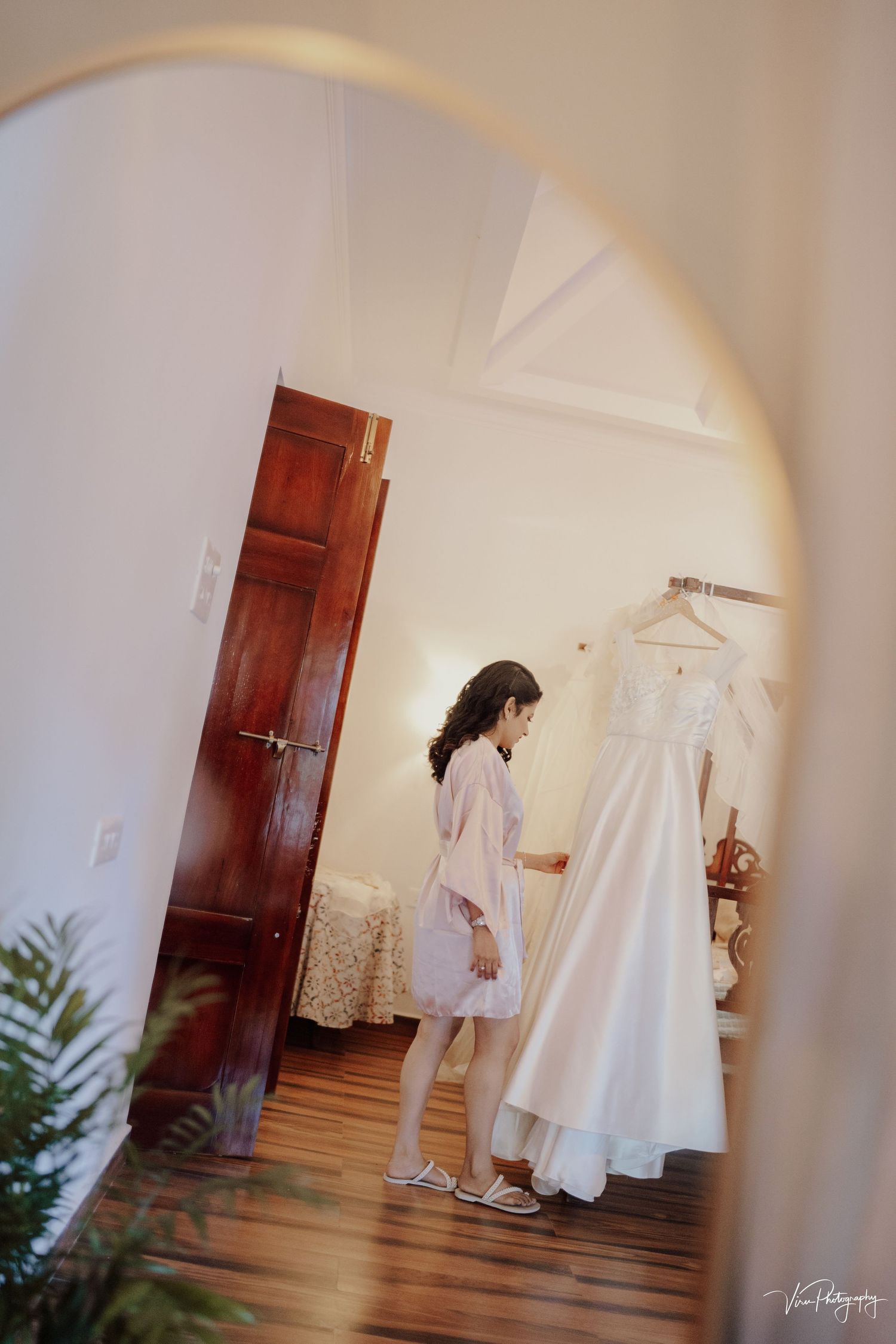 Through a doorway arch, soft light illuminates a bridal gown hanging in a room with hardwood floors and white walls.