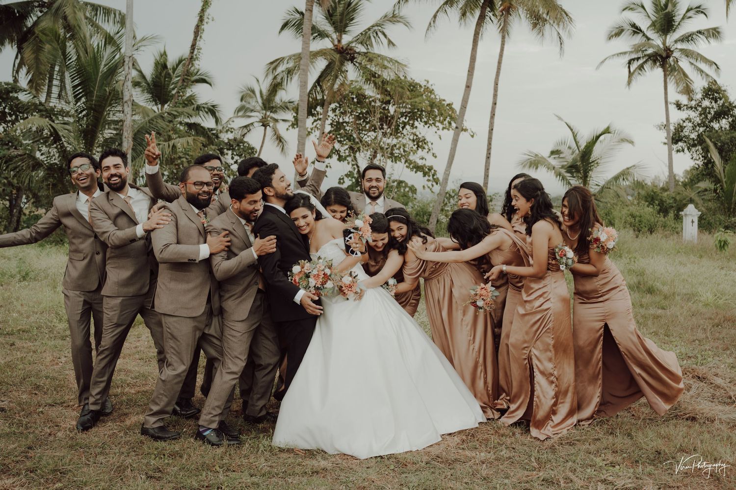 Wedding party poses together outdoors with palm trees in background wearing brown and rose gold attire.