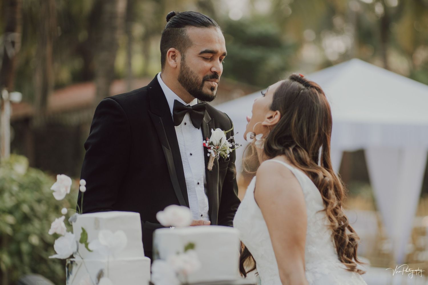 A joyful moment between newlyweds sharing a laugh next to their elegant white wedding cake outdoors.