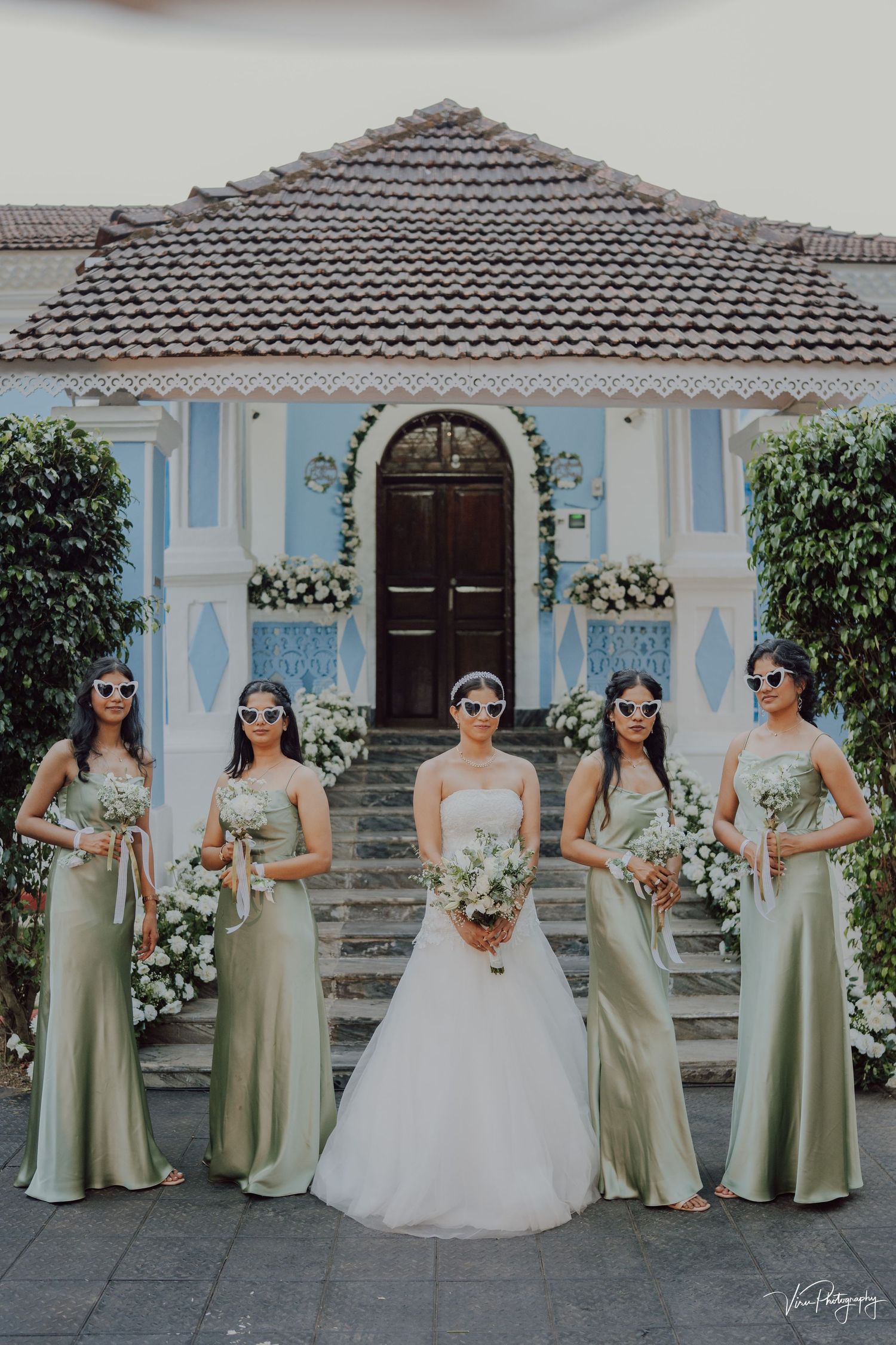 Bridesmaids in sage green dresses pose with bride in white gown outside elegant blue and white Mediterranean-style villa.