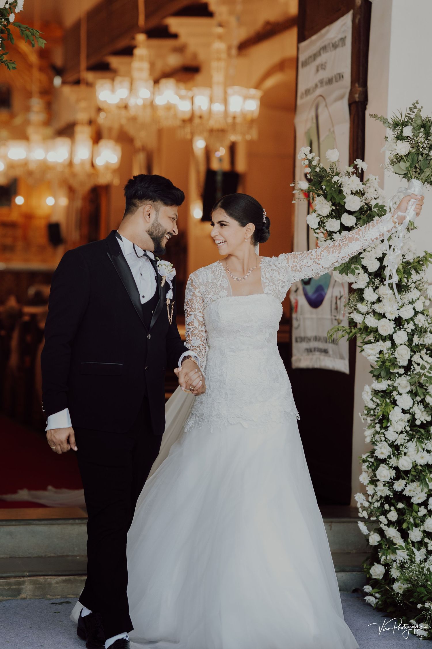 A bride in a long-sleeved lace wedding gown stands with her groom near elegant white floral arrangements and warm lighting.