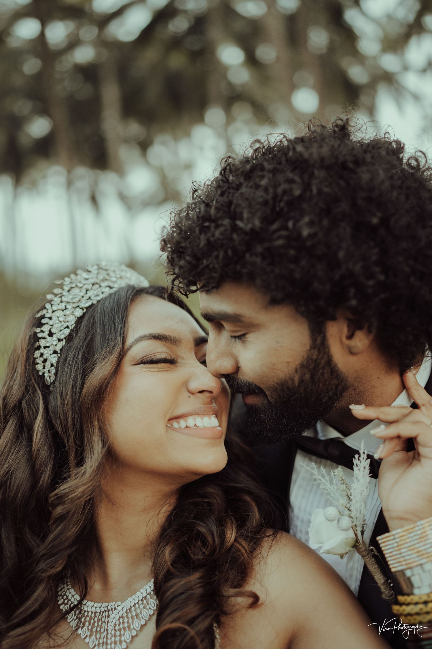 A romantic close-up portrait of a couple embracing outdoors, with the woman wearing a crystal headpiece and necklace.
