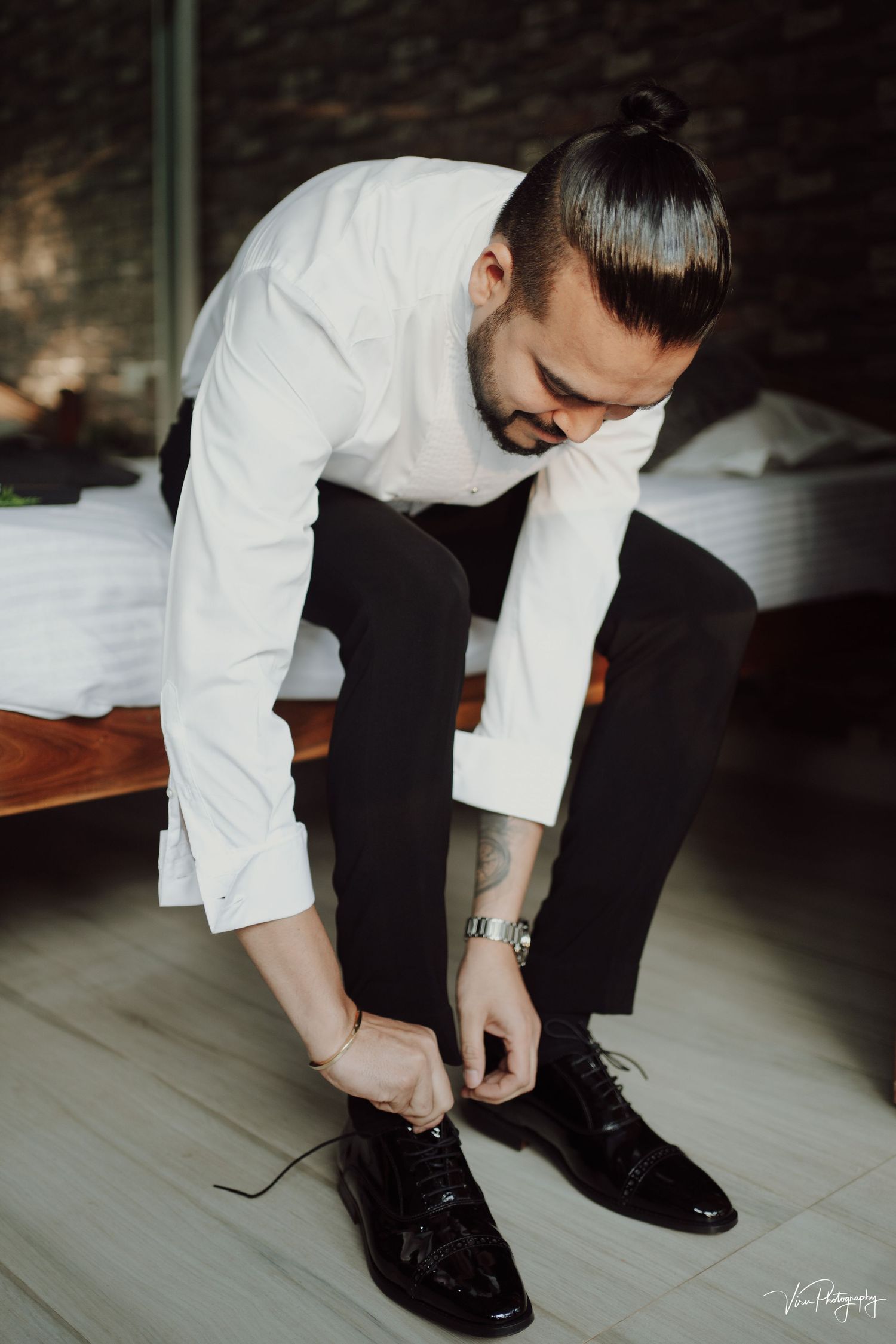 Someone in formal attire ties black dress shoes while getting ready on a wooden bench against a brick wall.