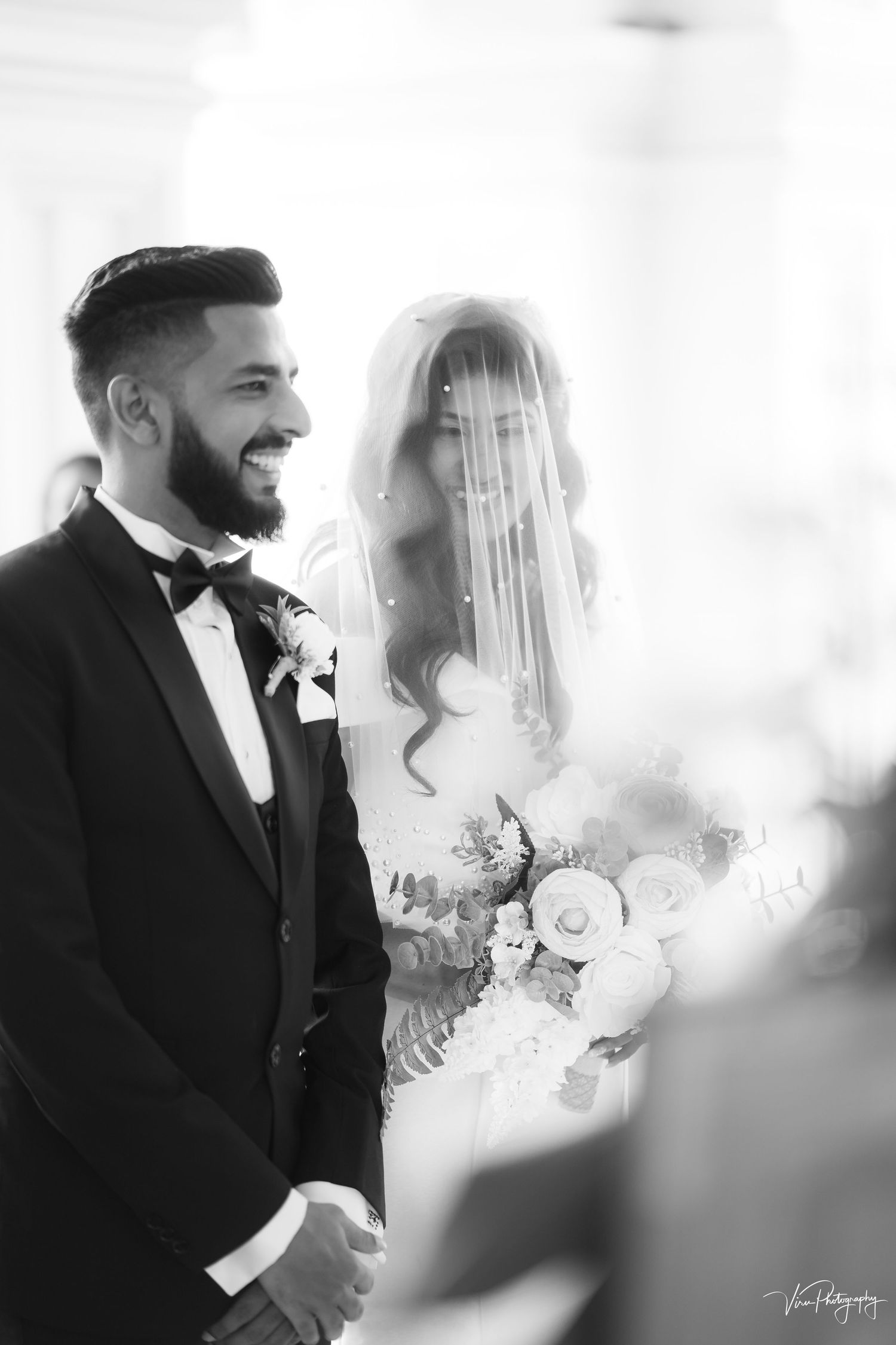 A black and white wedding photo with a bride in a veil holding flowers next to a bearded groom in a tuxedo.