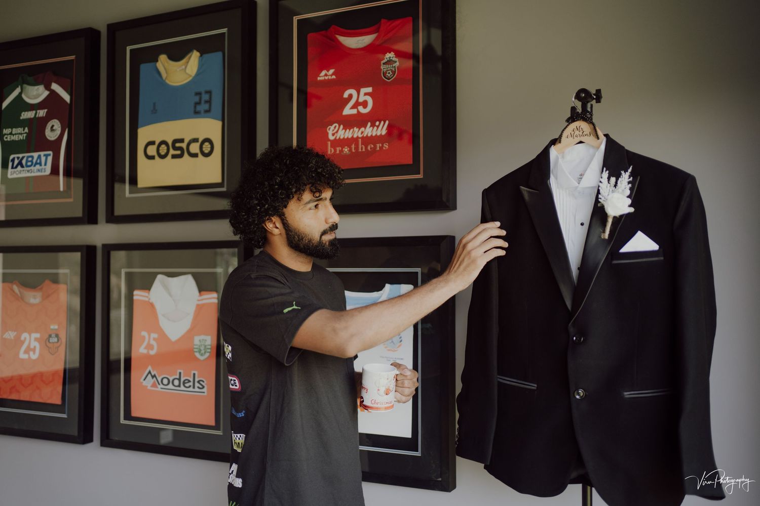 A person wearing a black t-shirt arranging a formal black tuxedo jacket displayed next to framed sports jerseys on a wall.