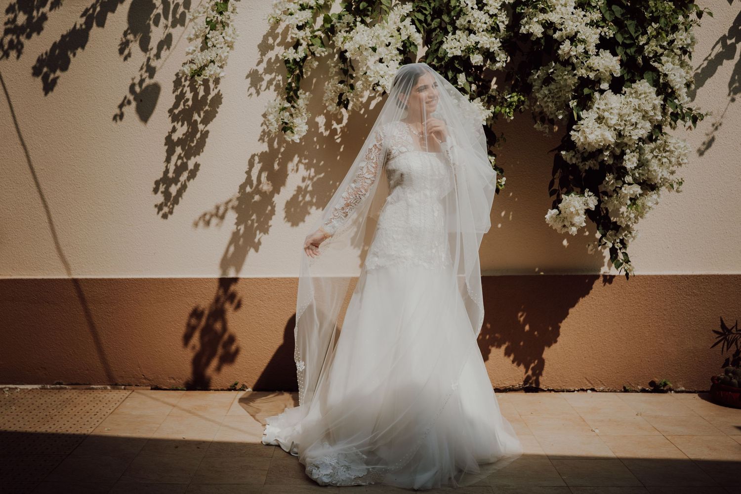 A bride in a flowing white wedding gown and long veil stands beneath hanging vines against a beige wall.