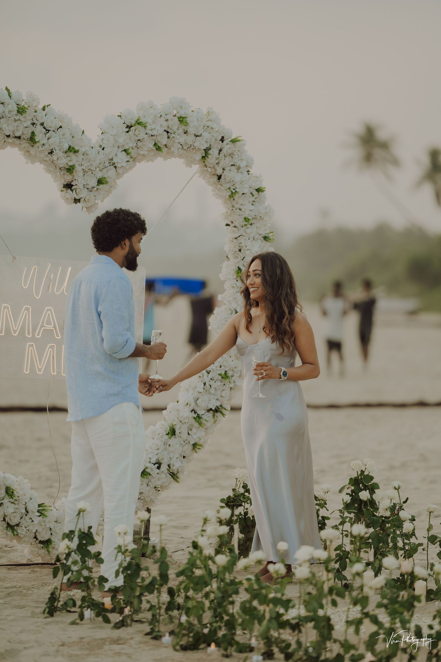 A romantic beach wedding ceremony setup with floral arch and couple in white outfits standing in the sand at sunset.