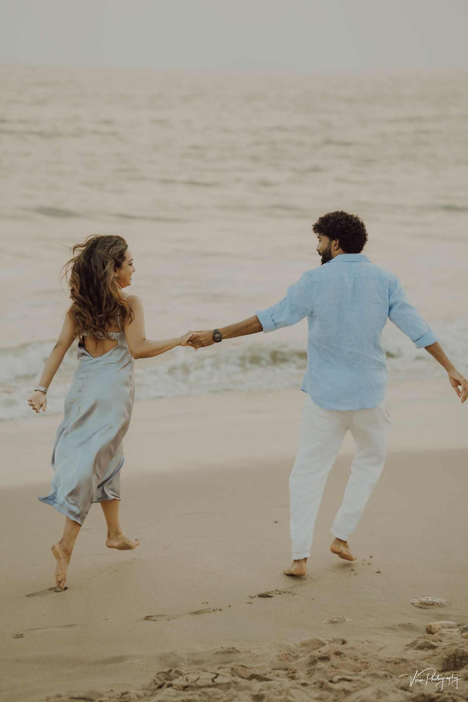 A couple holding hands while dancing together on a misty beach at sunset in coordinated blue and white outfits.