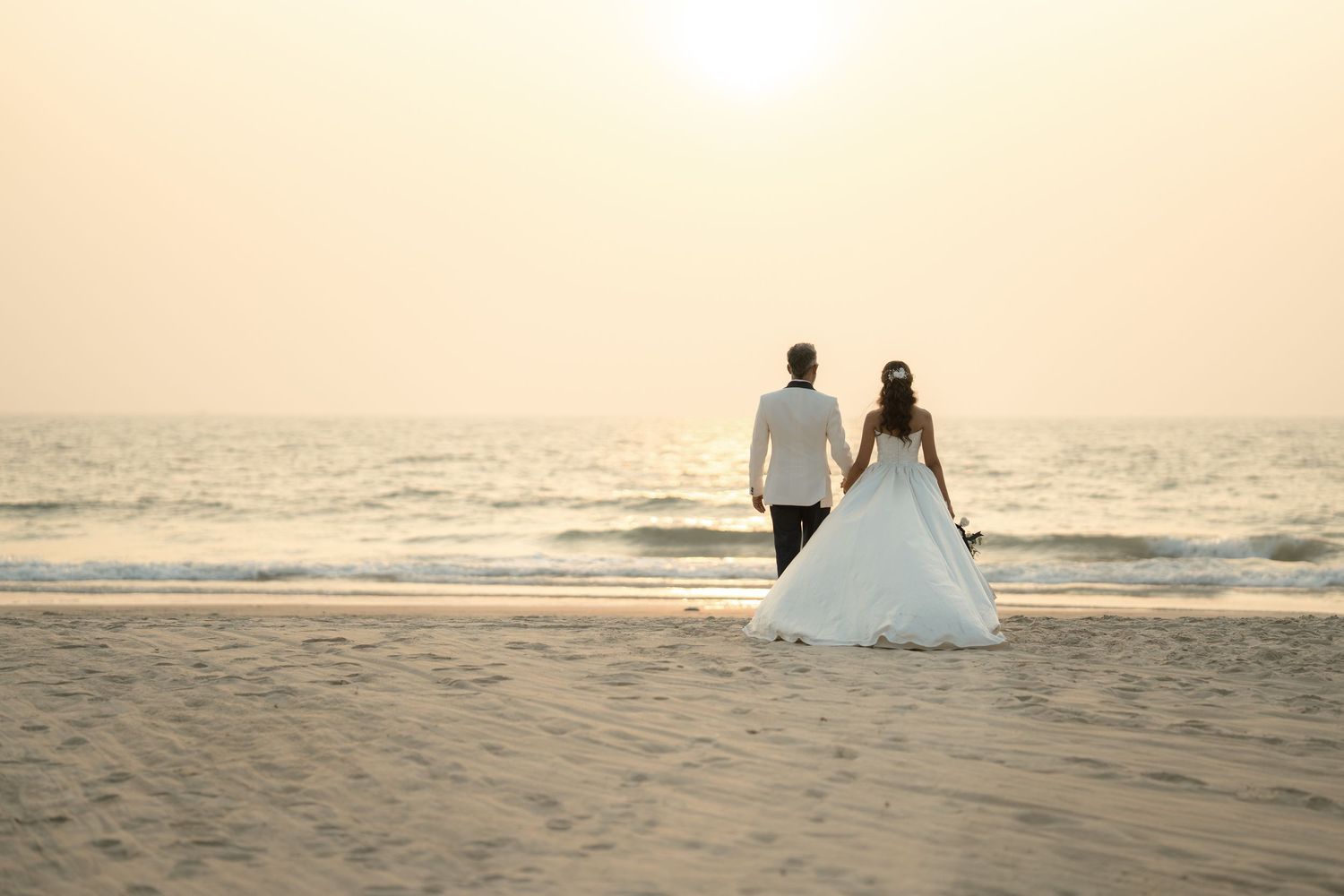 Bride and groom holding hands walking on a sandy beach at sunset with ocean waves in the background.