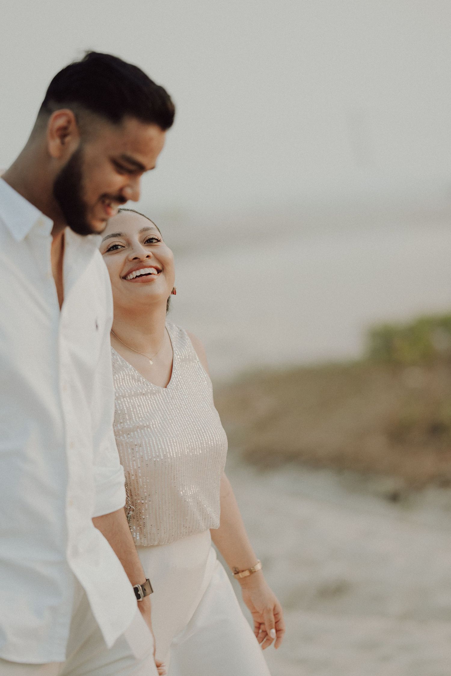 A smiling couple in white outfits share a romantic moment on a beach, with the woman wearing a hijab.