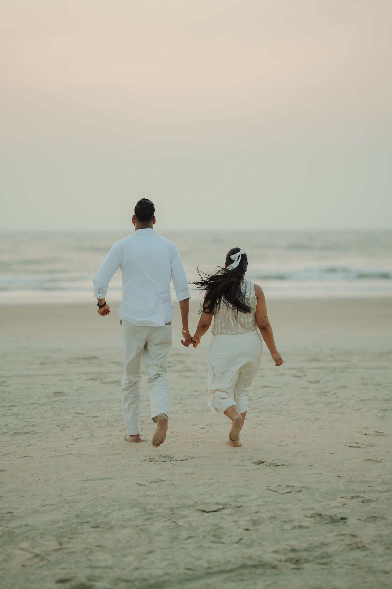 Couple in white outfits holding hands running barefoot on a serene beach at dusk.