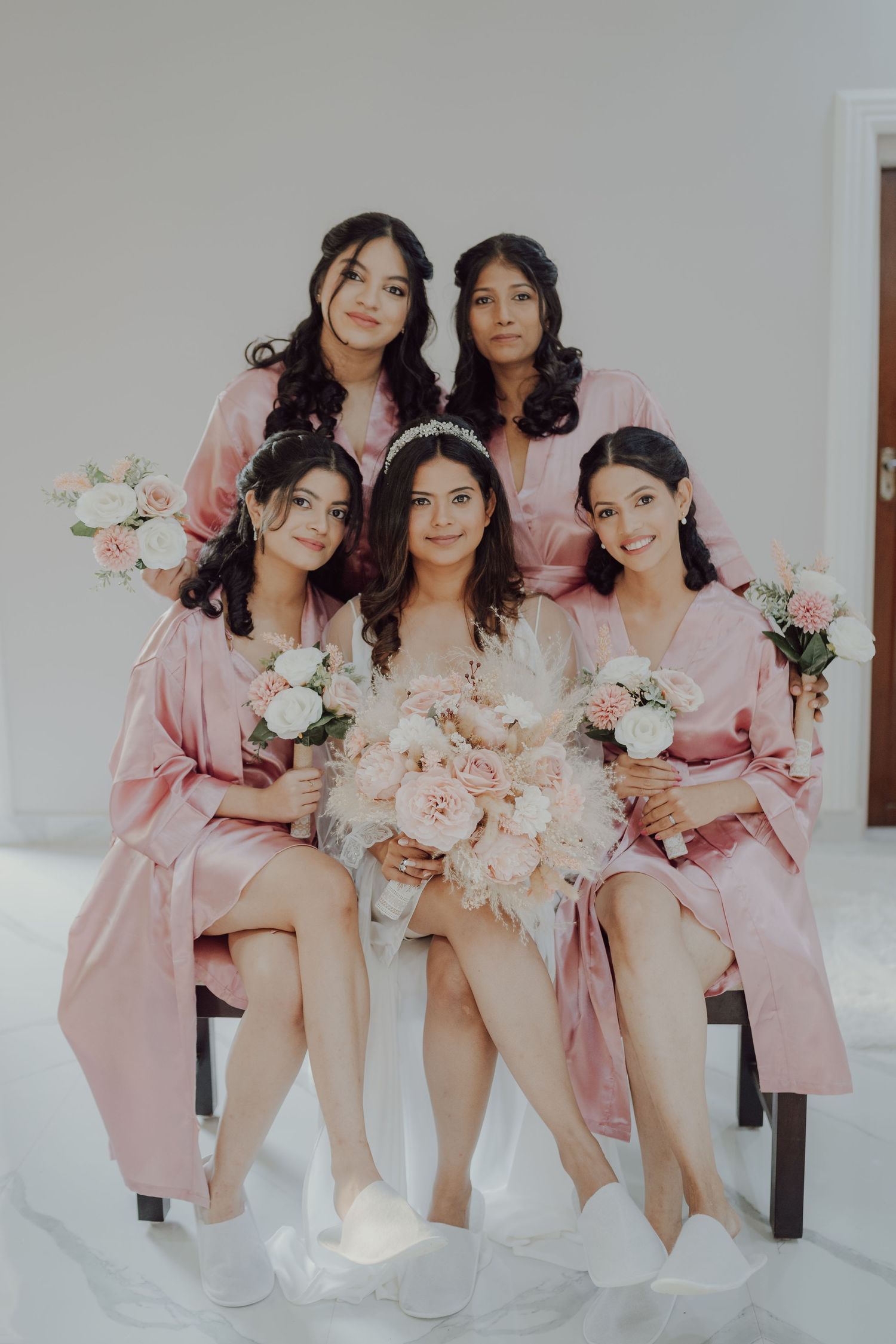 Bridal party in matching pink robes holding floral bouquets pose together.