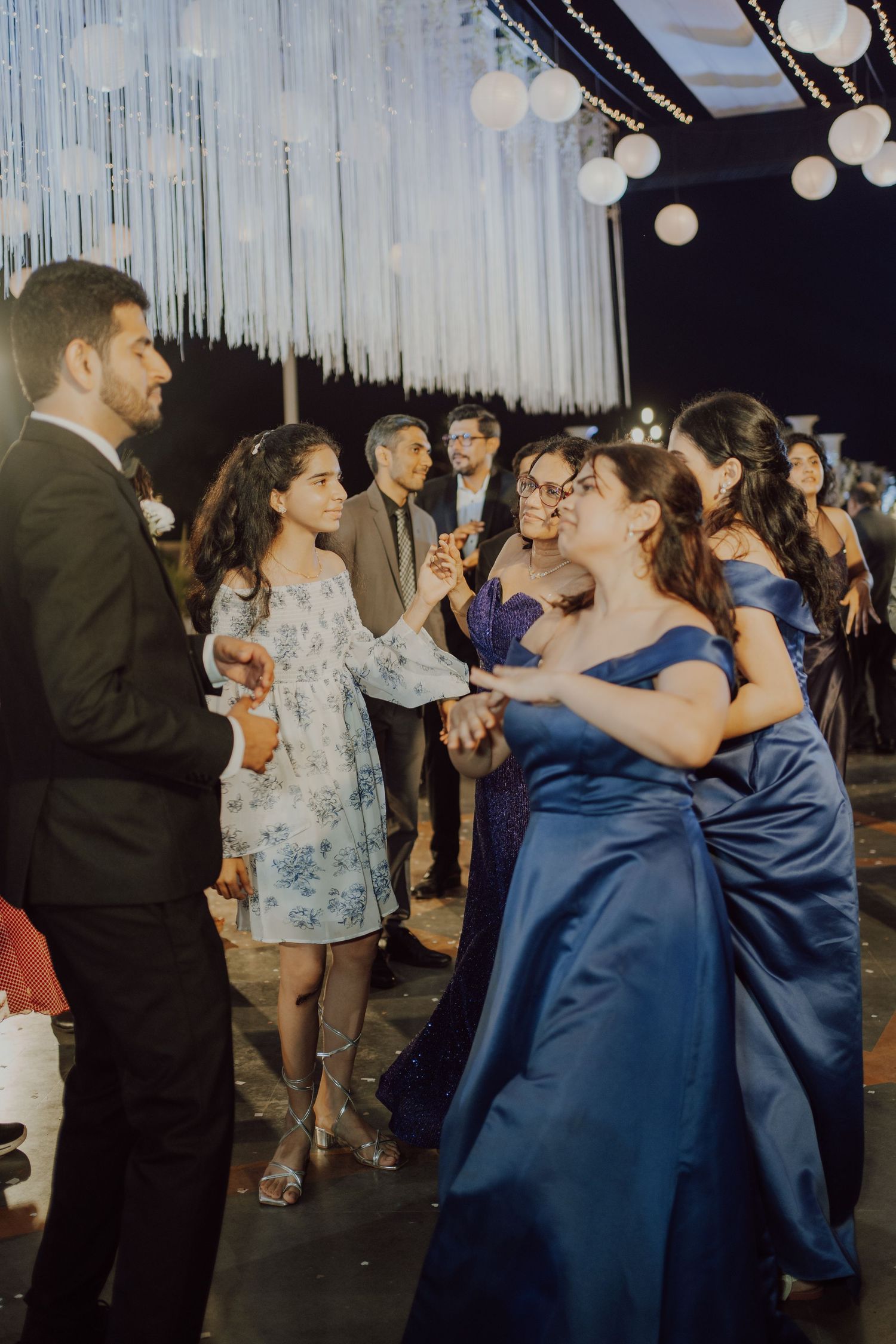 Wedding party dances under white streamers and glowing orb lights during evening celebration.
