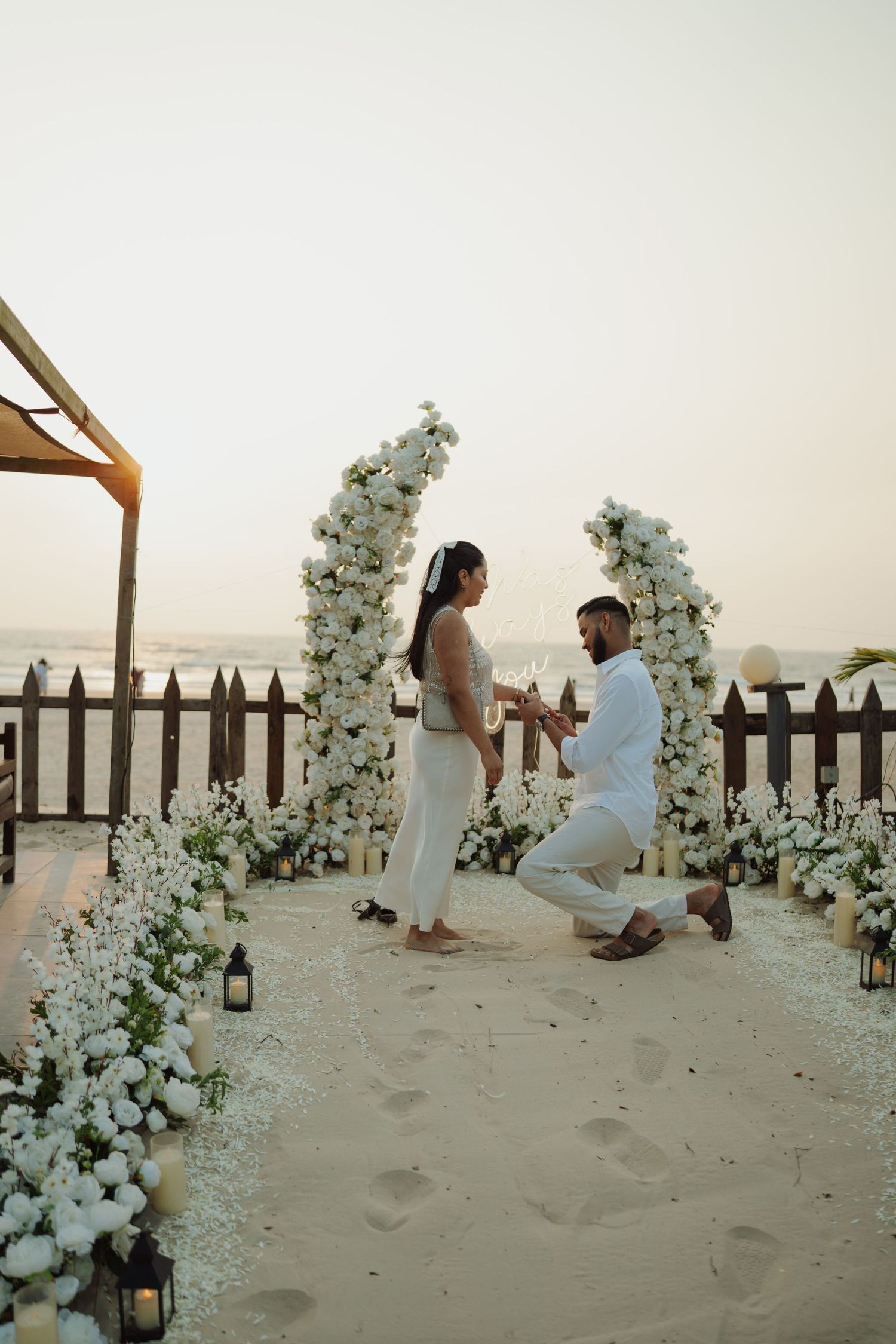 Man proposes to woman on one knee on sandy beach surrounded by white floral arrangements at sunset.