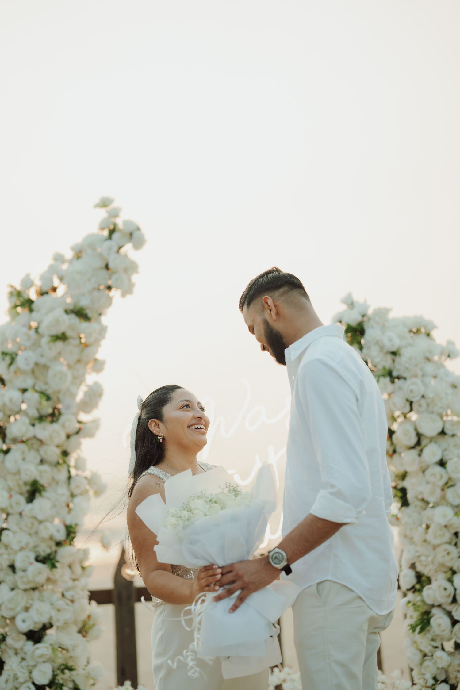 Couple in white attire exchanging vows surrounded by white floral arrangements at an outdoor wedding ceremony.