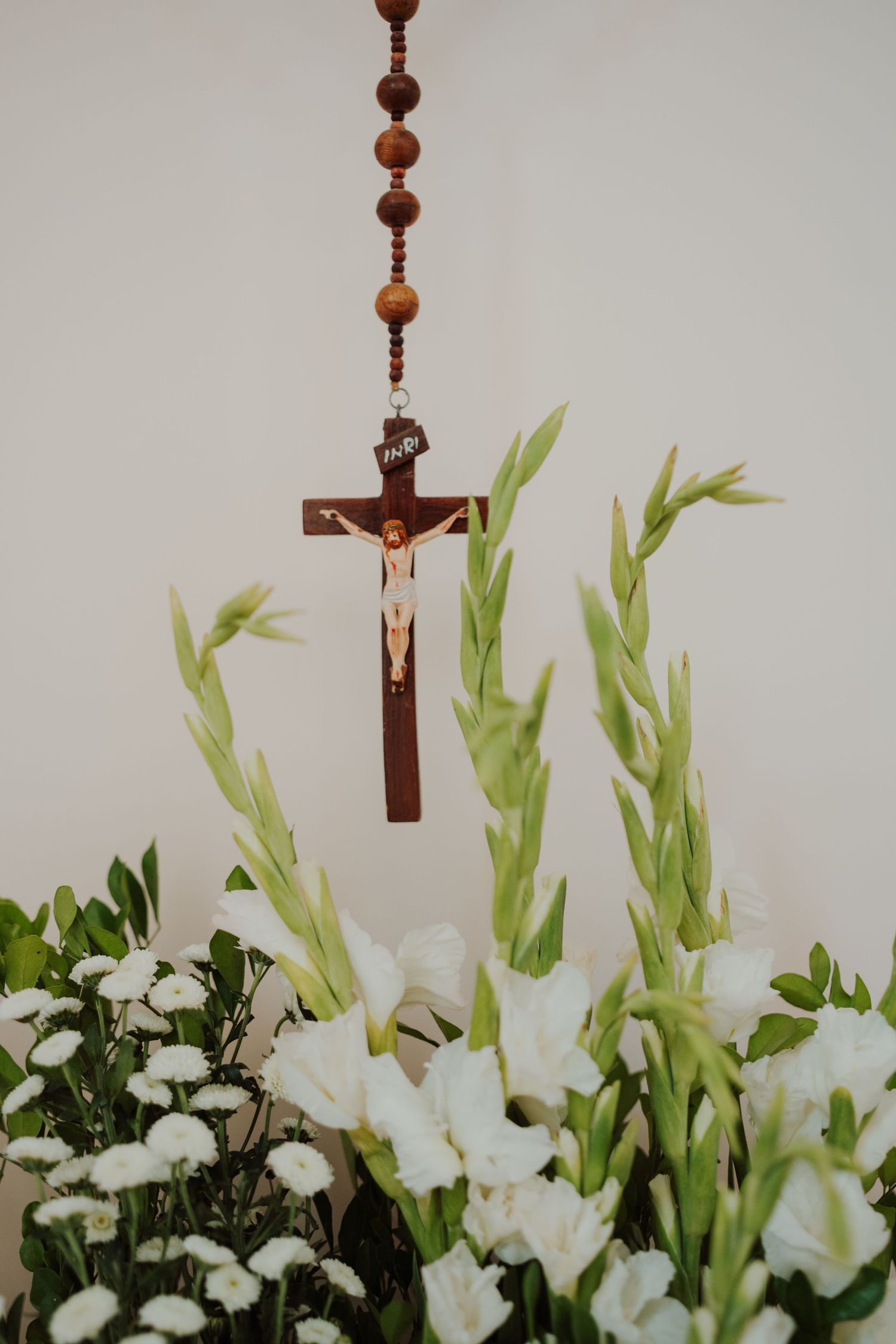 Wooden rosary and crucifix hang above a lush arrangement of white gladiolus flowers and green foliage against a plain wall.