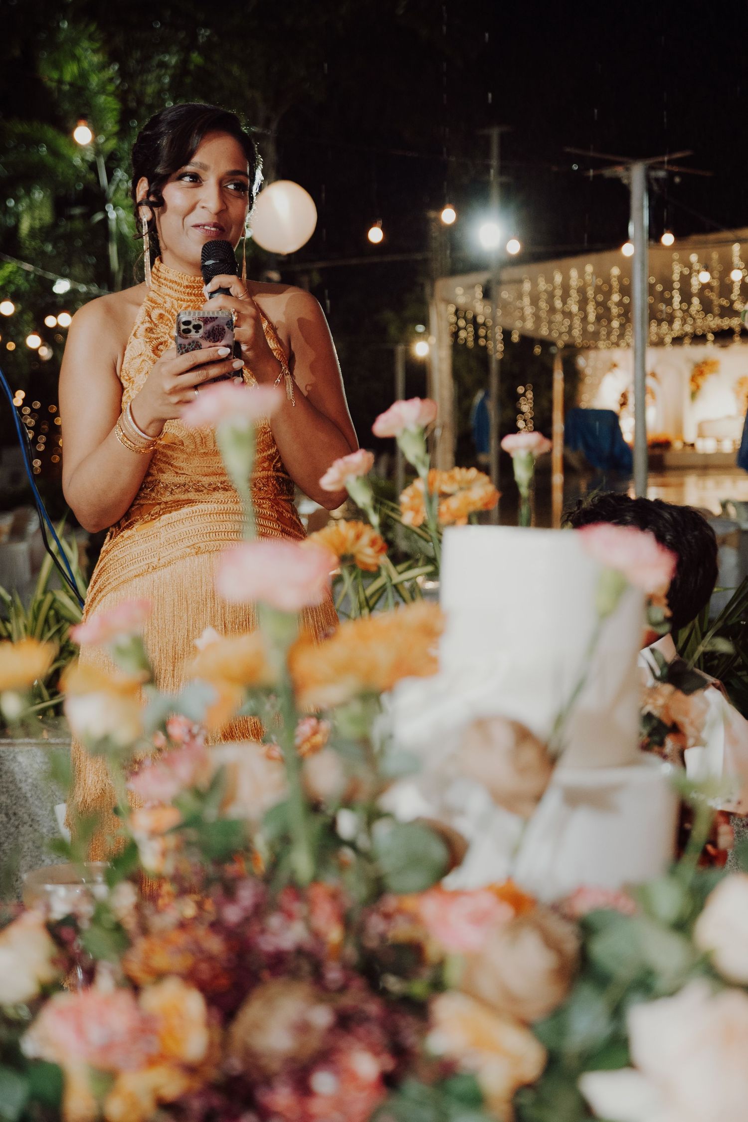 Woman in a golden fringe dress speaks into a microphone at an evening wedding reception behind a floral centerpiece and white cake.