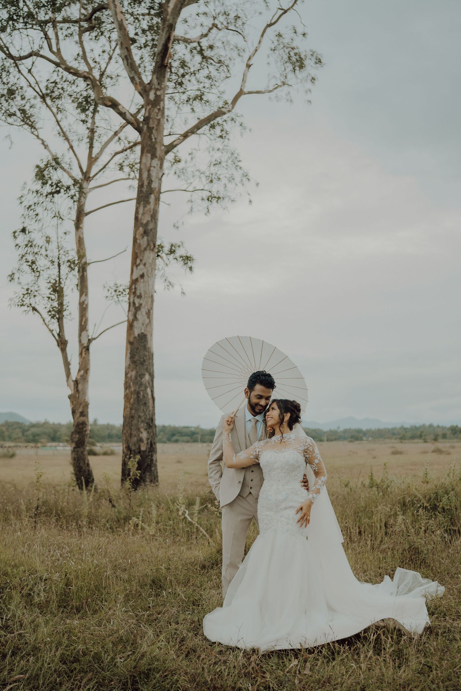 Bride in a lace wedding gown and groom in a light suit embrace beneath a white parasol in a grassy field with tall eucalyptus trees.
