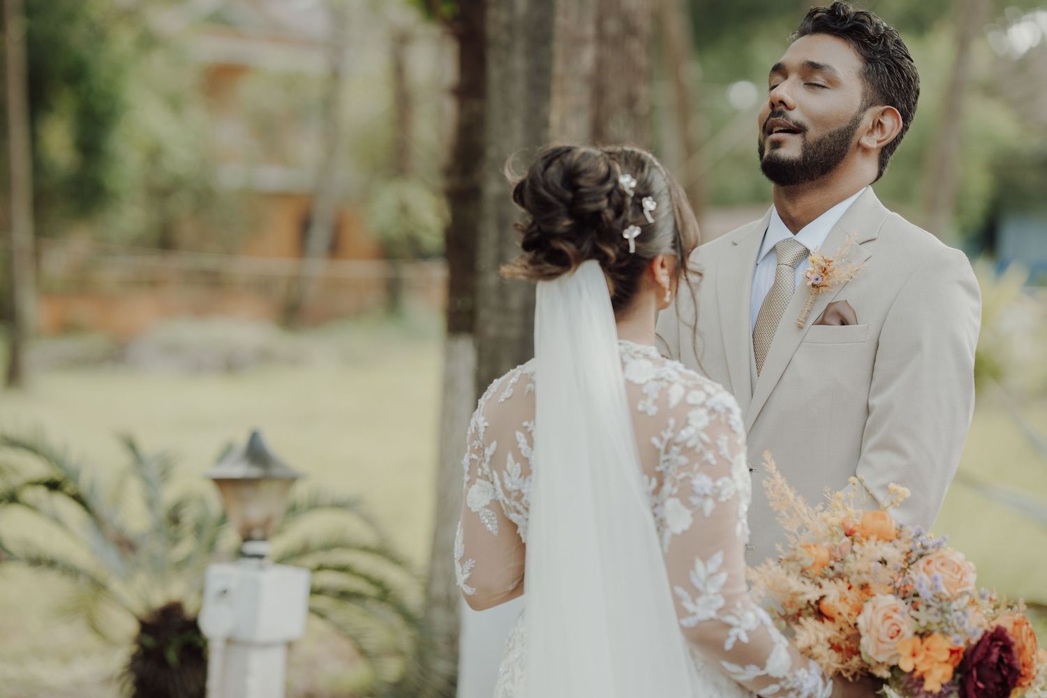 A groom in a beige suit smiles at his bride in a lace gown and veil as she holds a peach and orange bouquet outdoors.